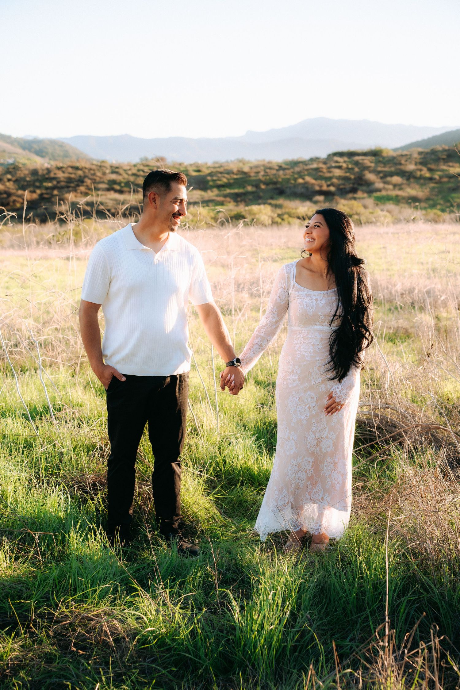 Couple holding hands in the grass with mountains behind, captured in a timeless, in-the-moment engagement session.