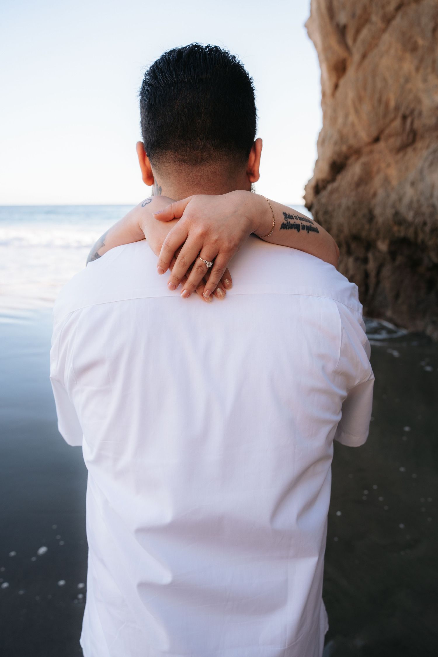 Close-up view of hands gently resting on shoulders in white clothing against ocean backdrop.