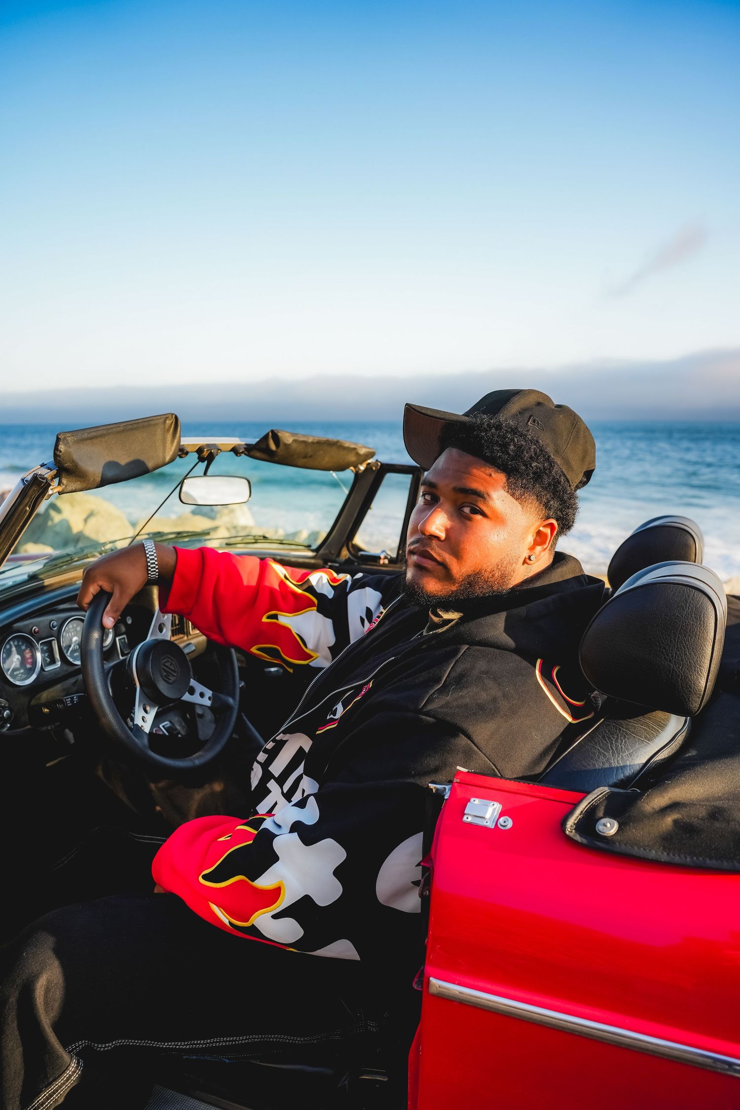Person driving a red convertible car along coastal road at sunset.
