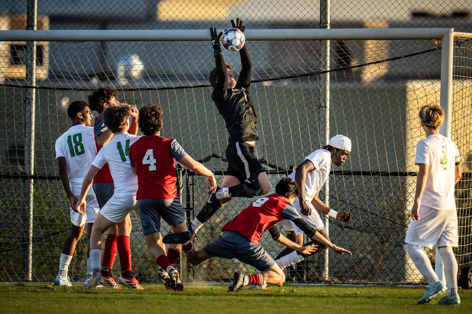Soccer goalkeeper leaps to catch ball during competitive match with players in green, white, and red uniforms.