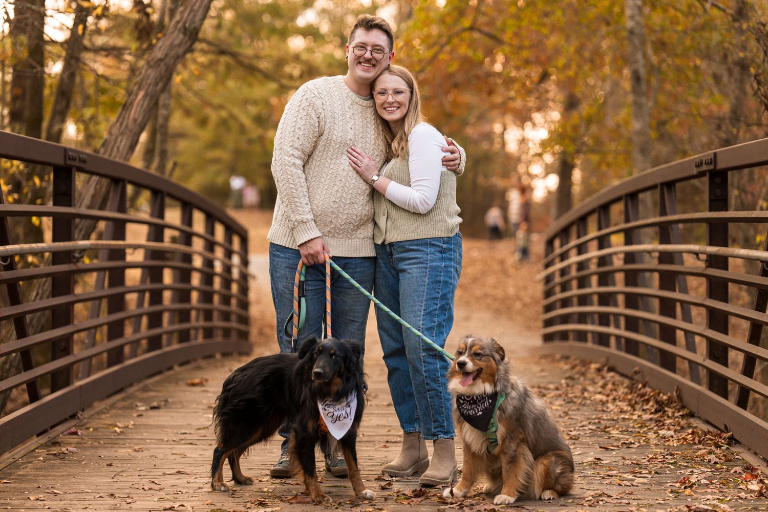 A couple and their black dog walk together on a wooden bridge surrounded by autumn foliage.
