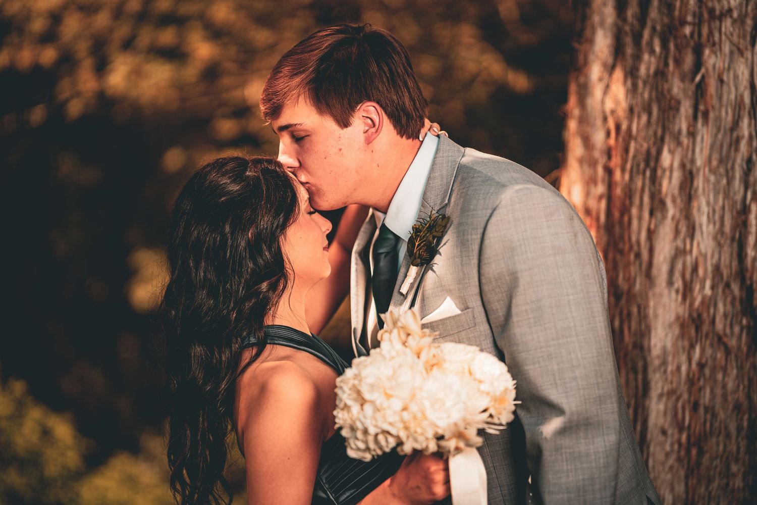 A bride and groom share a romantic kiss outdoors near a tree, with the bride holding a white bouquet on their wedding day.