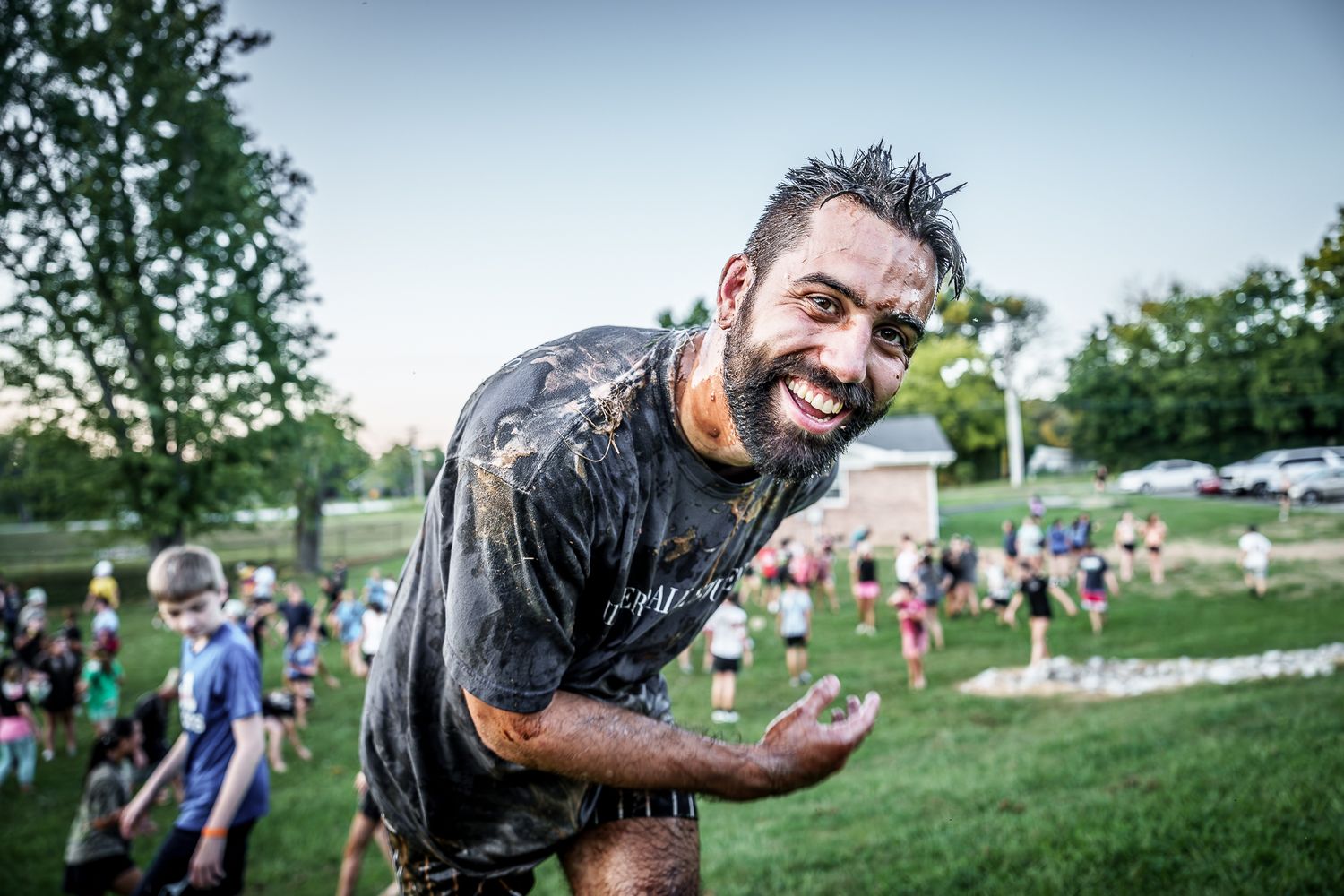 Smiling muddy man covered in dirt at outdoor mud run event with crowd and green trees in background.