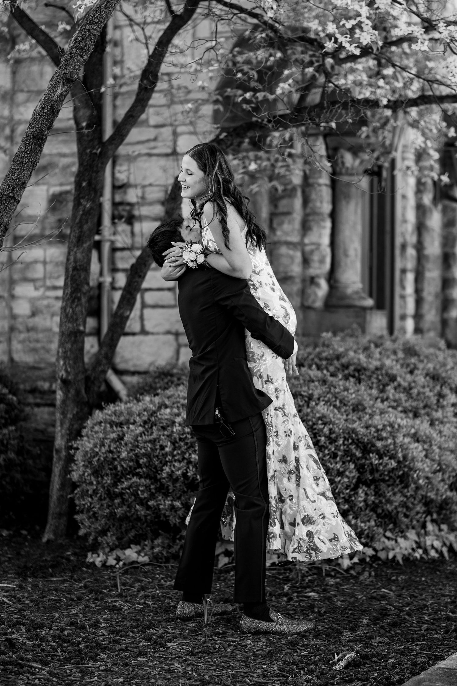 Black and white portrait of couple embracing near stone building and landscaping.