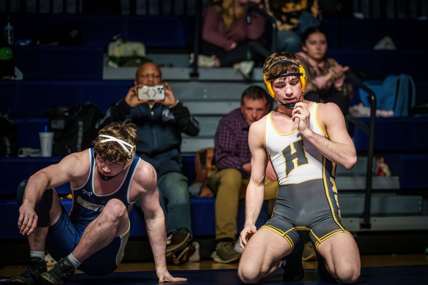 Two wrestlers compete on a mat during a match while spectators watch from the bleachers in the background.