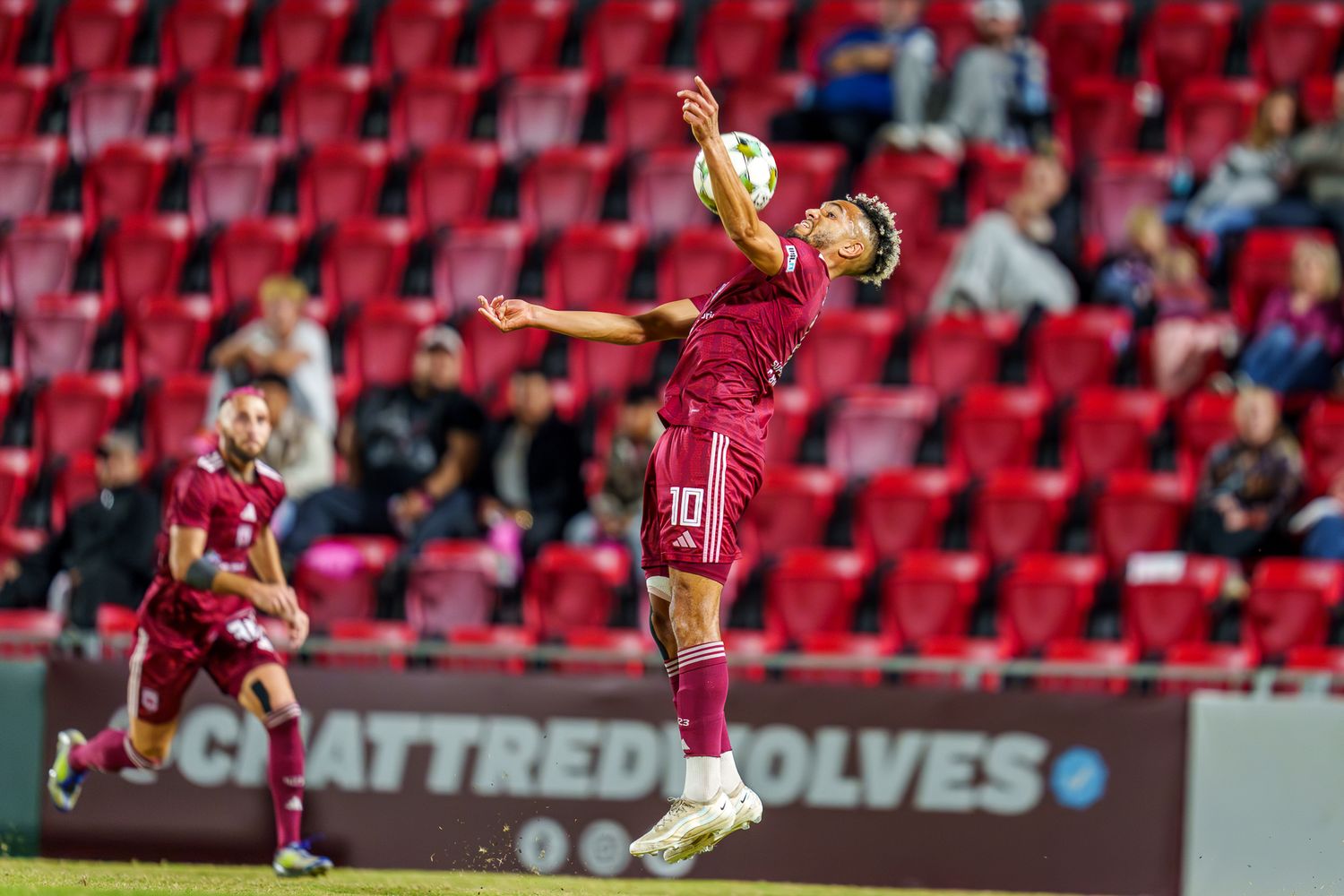 Soccer player jumps high to control the ball against red stadium seating backdrop.