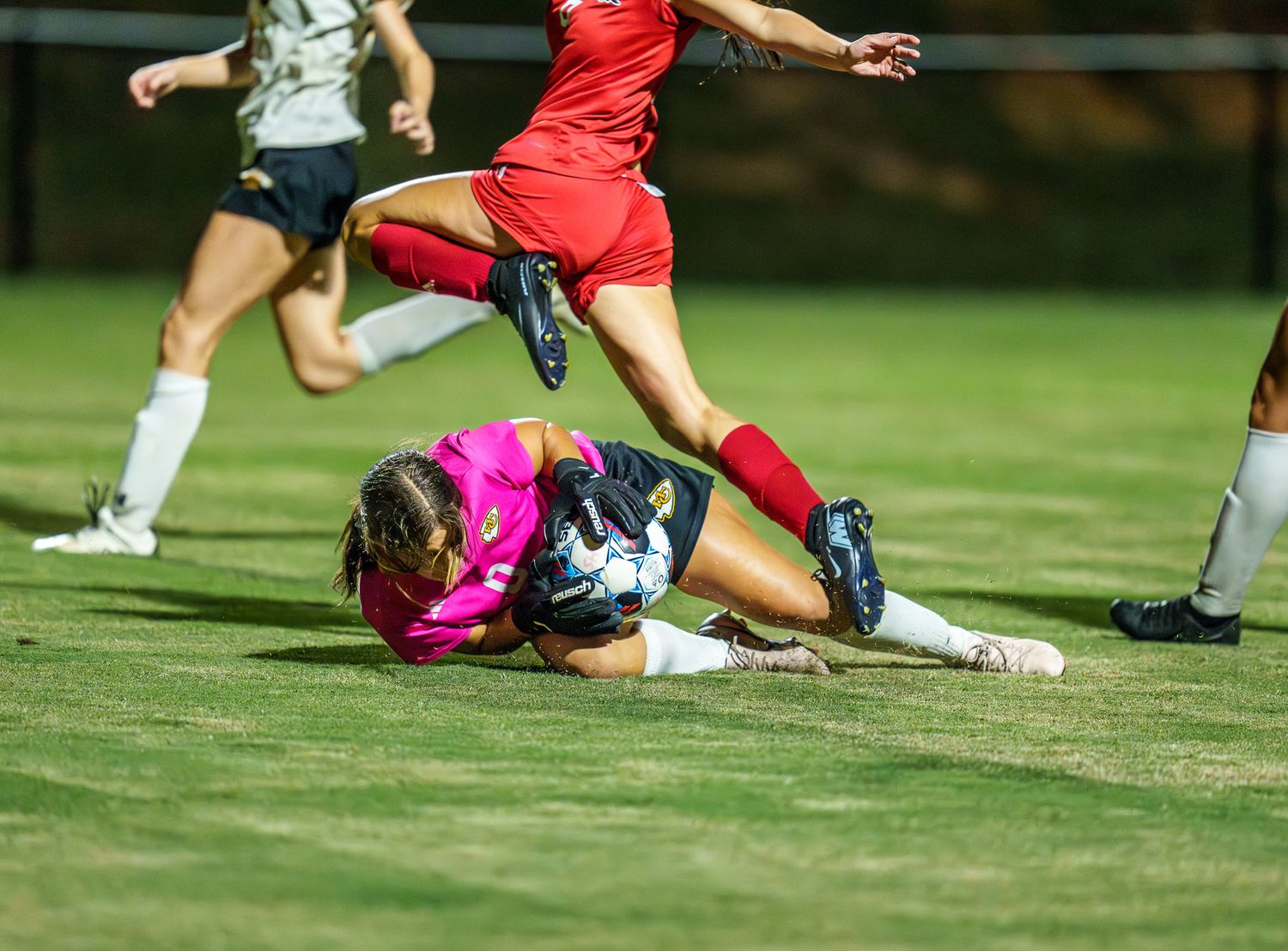 A goalkeeper in pink dives to save the ball as a red-uniformed soccer player leaps over during a night match.