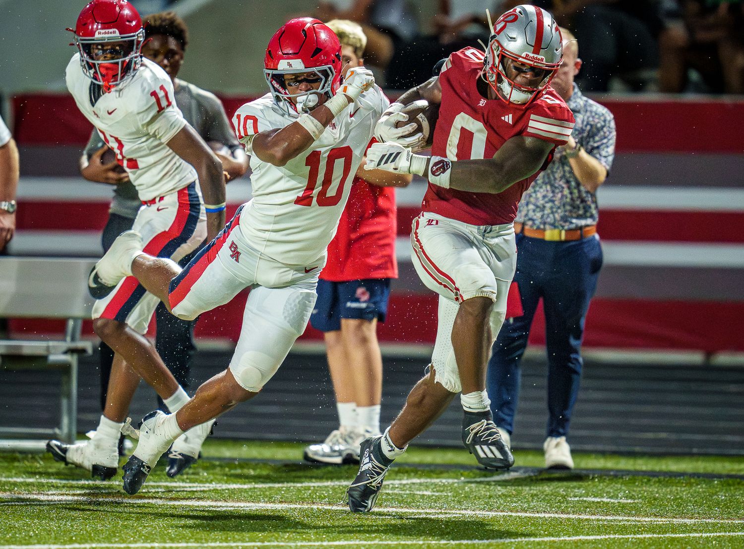 Two football players in red and white uniforms compete during a high school football game on a red turf field.