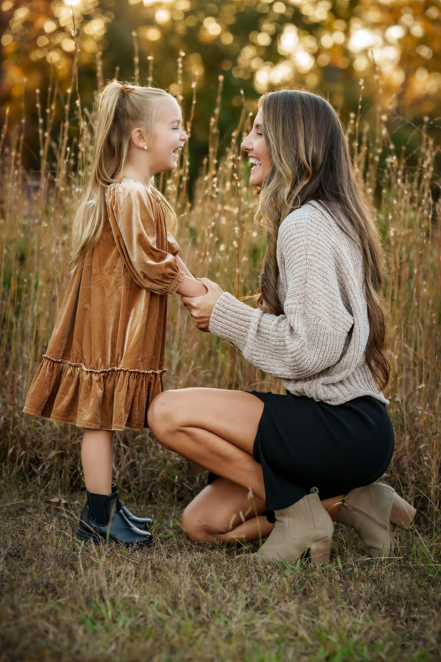 A mother kneels in a golden field, smiling and holding hands with her young daughter in a brown velvet dress at sunset.