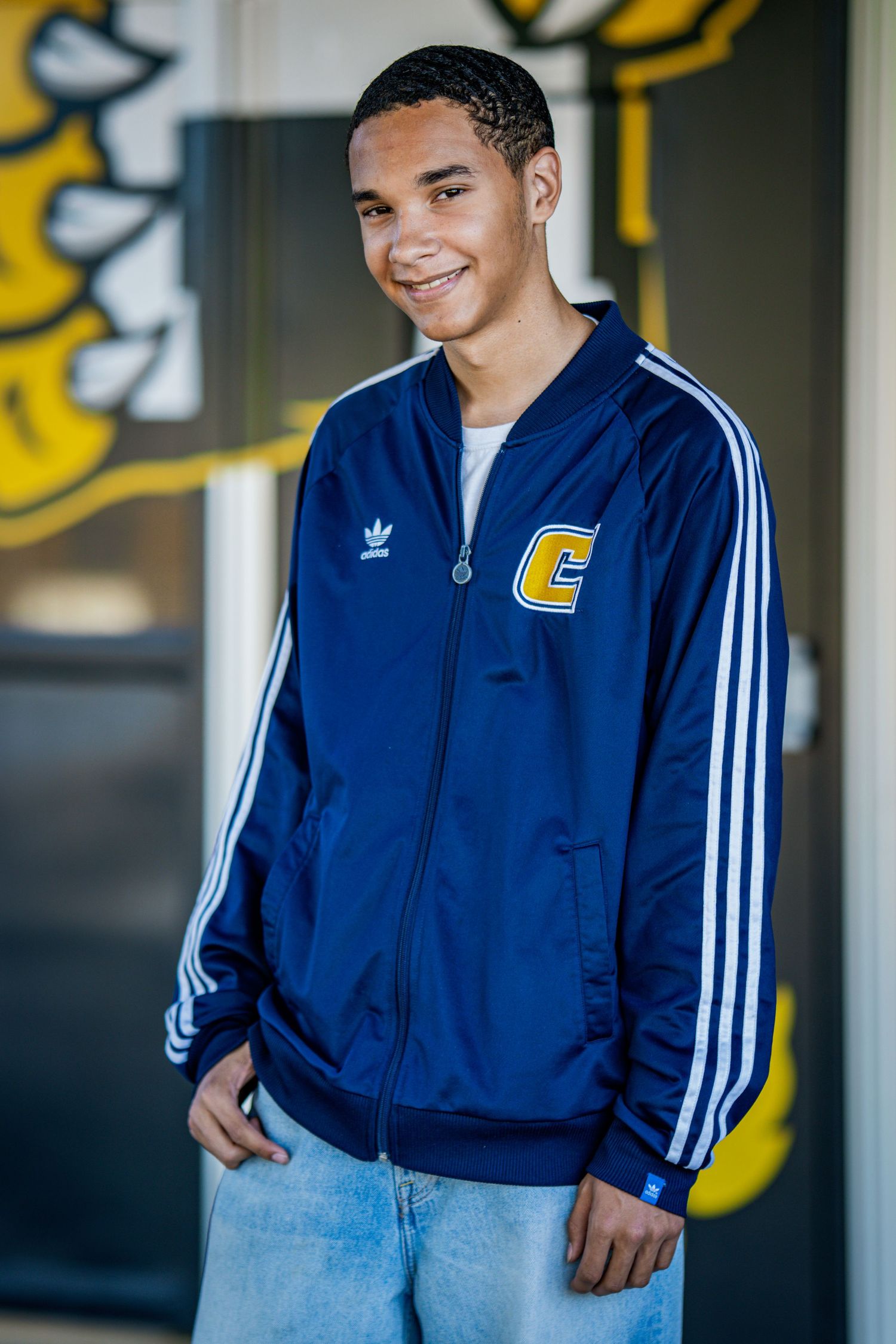 Student wearing blue Adidas track jacket posing against school mascot background.