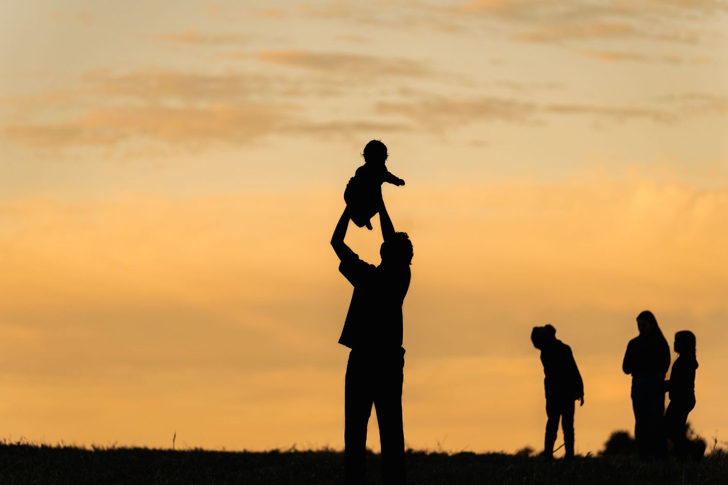 Silhouette of a father lifting a baby into the air at sunset while three family members stand nearby against a golden orange sky.