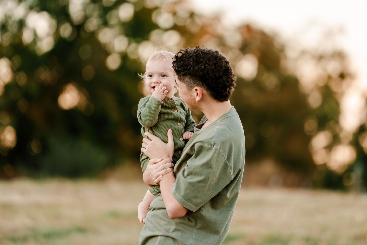 Man in an olive green shirt holds a baby in a matching green outfit while standing in a sunlit grassy field with softly blurred trees.