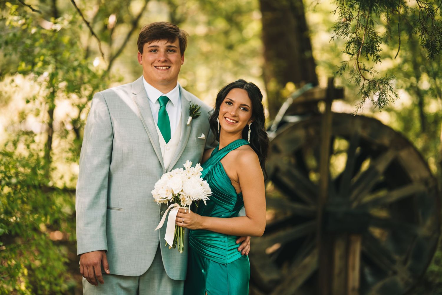 A couple in matching emerald green formal wear poses outdoors near a wooden wagon wheel surrounded by lush trees.