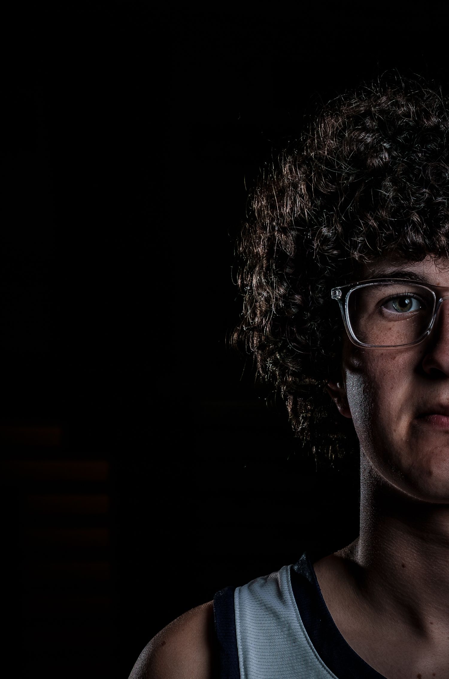 A young person with curly hair and glasses poses dramatically in low-key studio lighting against a dark background.