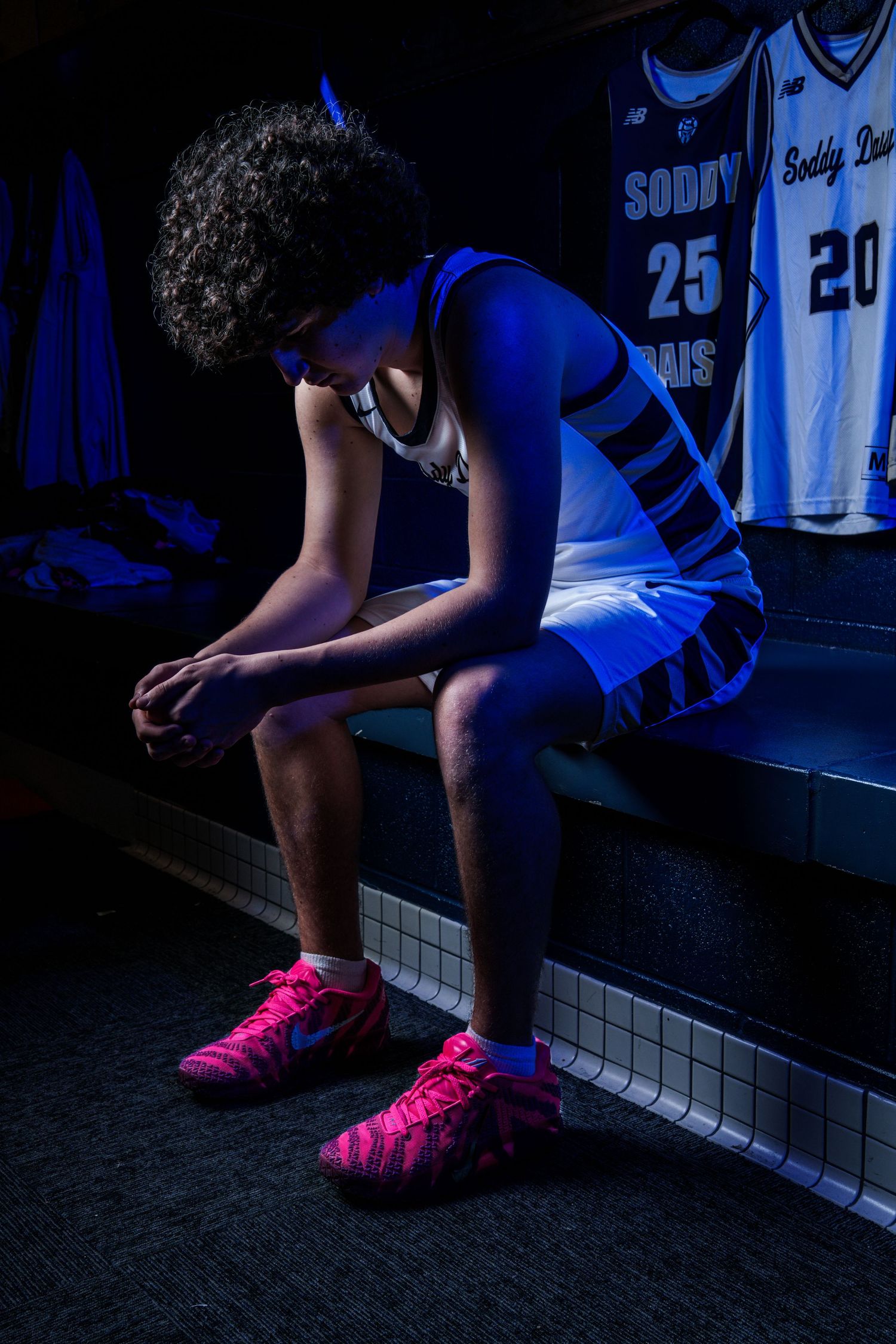 Basketball player wearing bright pink sneakers sits on bench in dark locker room with blue jerseys visible in background.