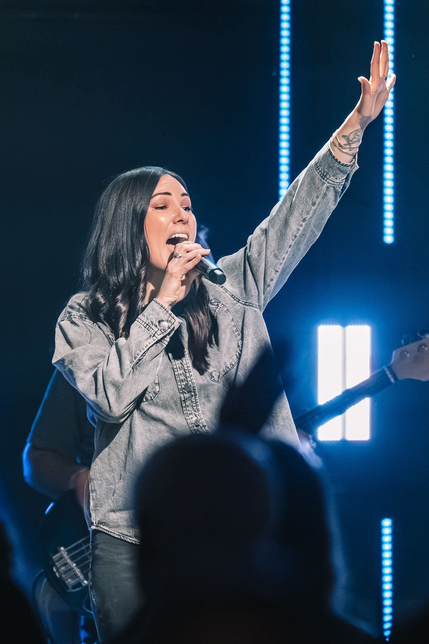A female singer performs on stage, raising one hand while holding a microphone, lit by blue stage lights.