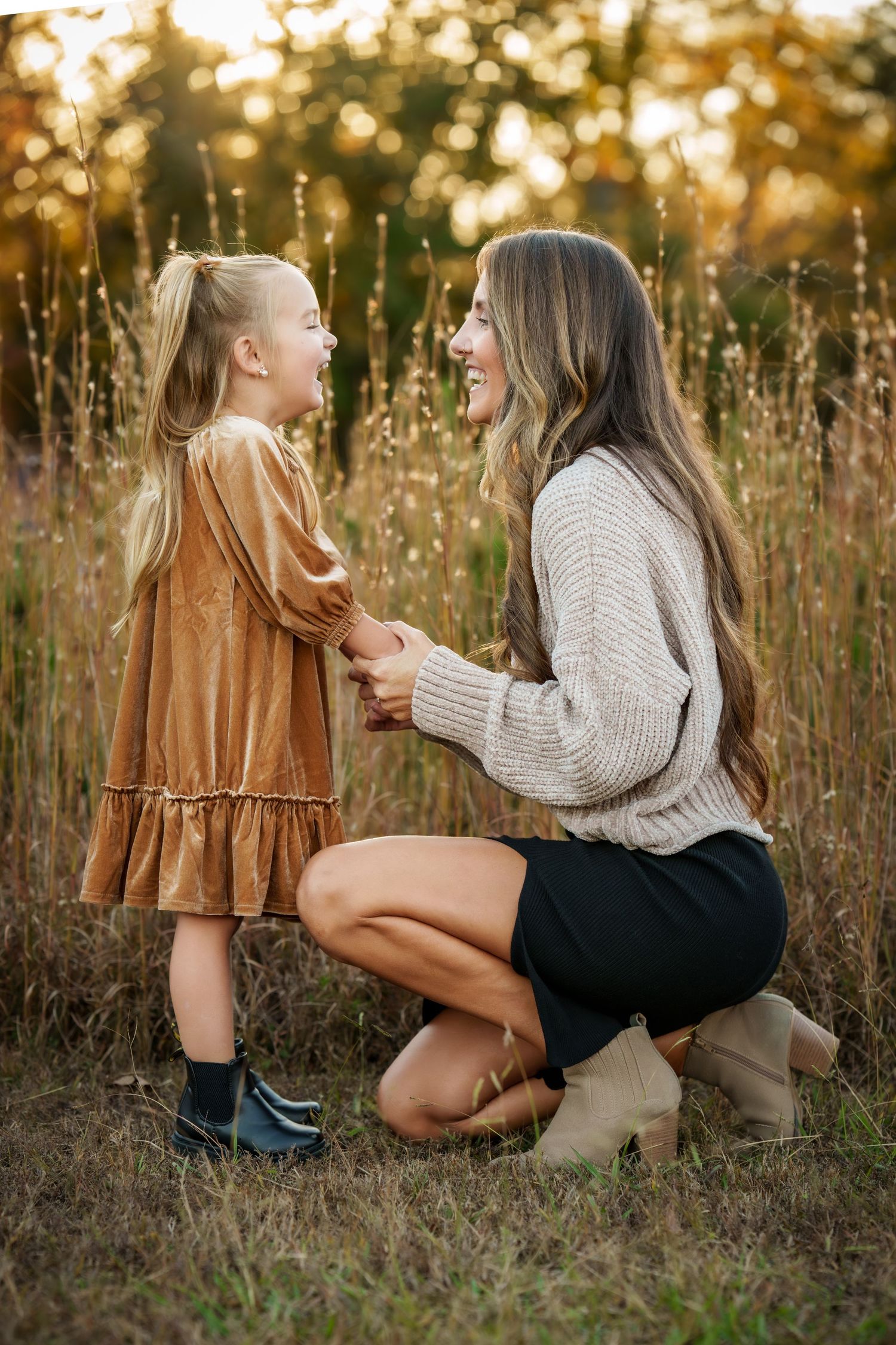 A mother kneels in a golden field, smiling and holding hands with her laughing young daughter at sunset.