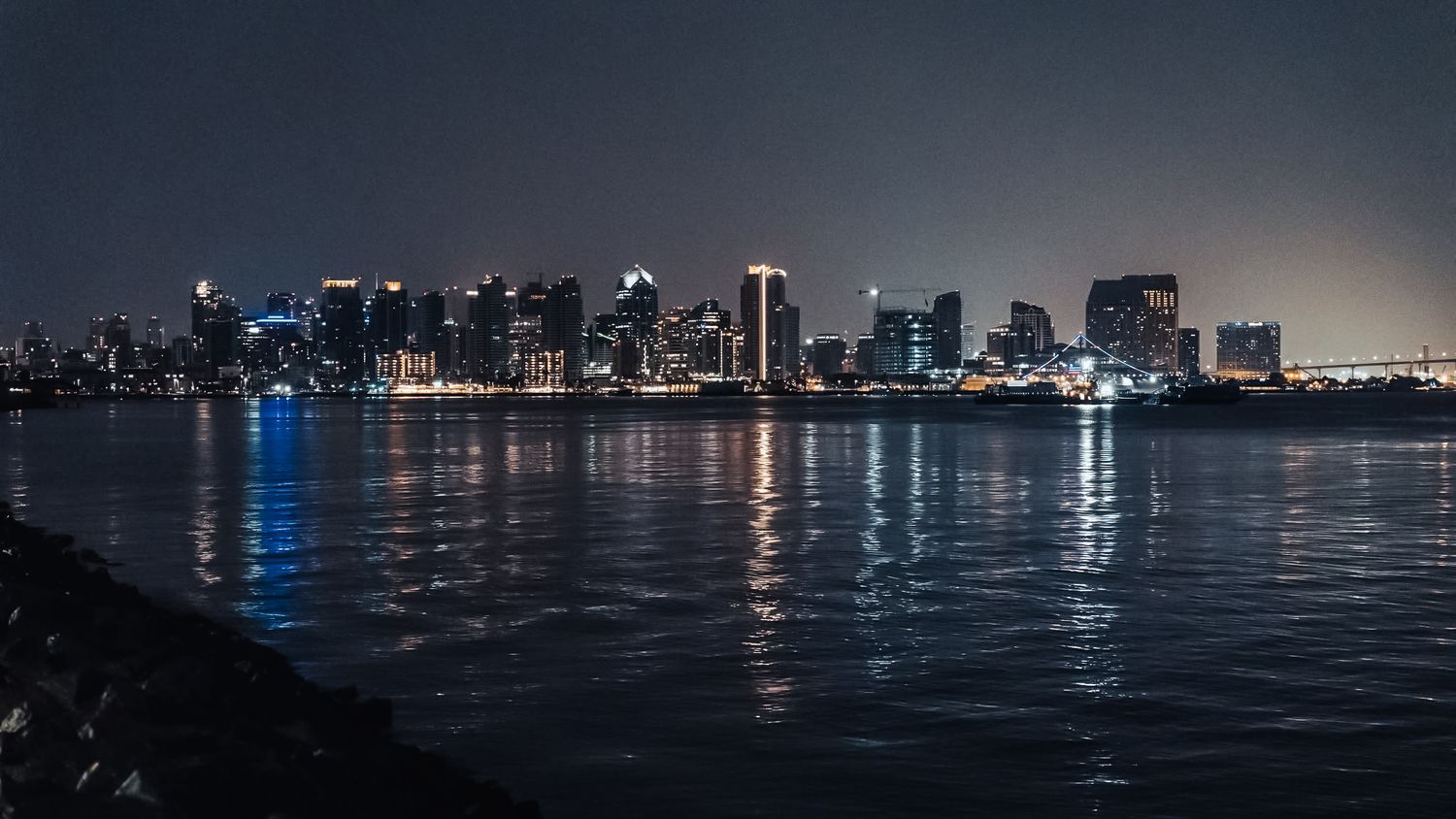 City skyline at night with illuminated buildings reflecting on dark water, creating a moody urban waterfront scene.