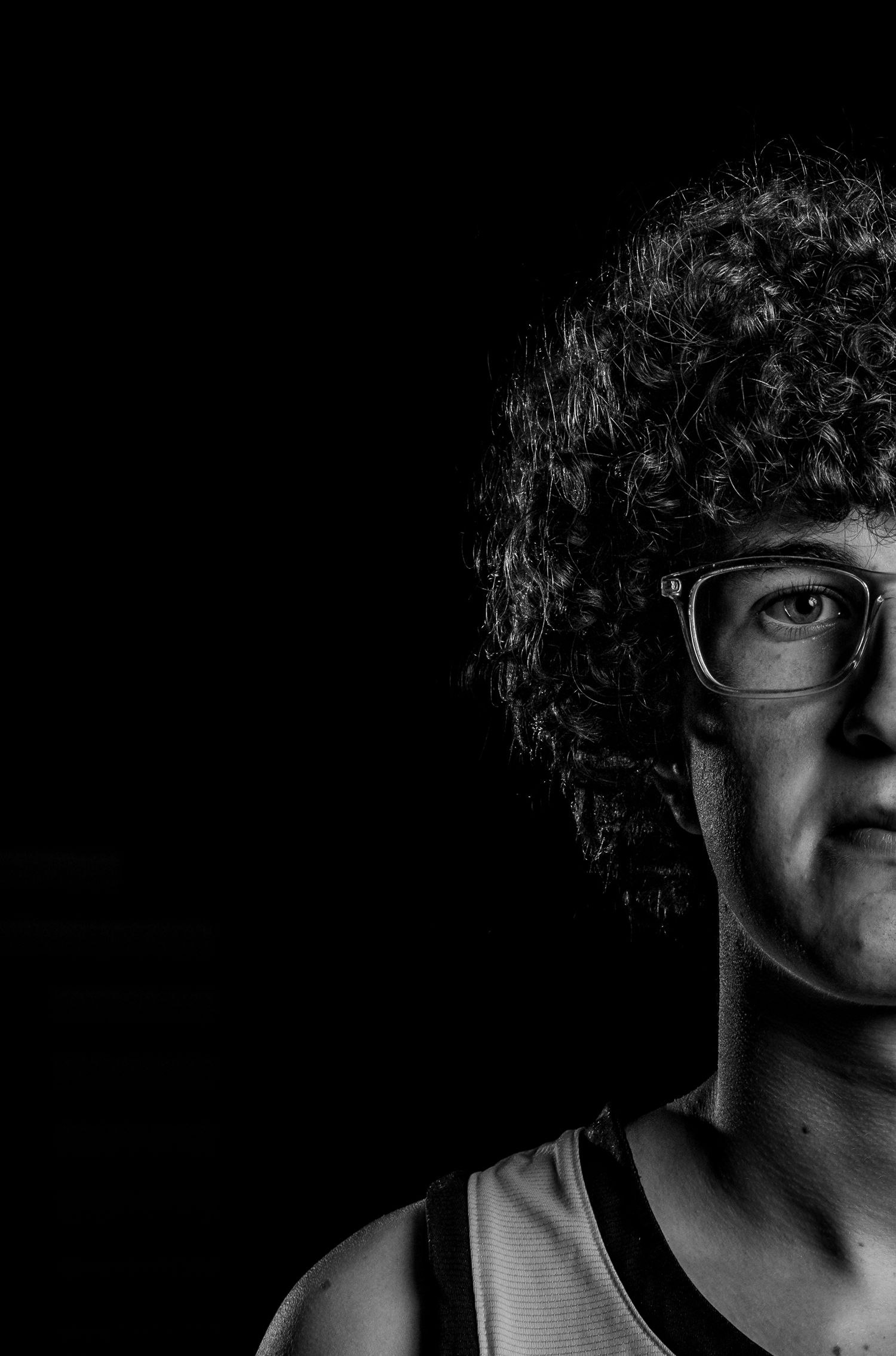 Black and white portrait of a curly-haired person wearing glasses, half face in shadow against dark background.