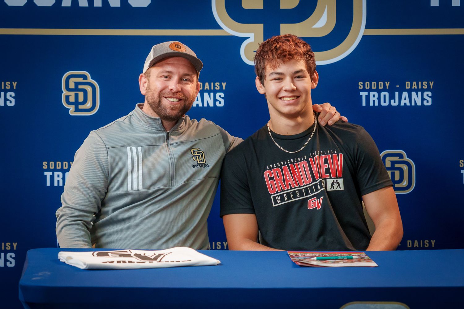 Two young men smile together at a Soddy Daisy Trojans signing day event, one wearing a Grand View University shirt.