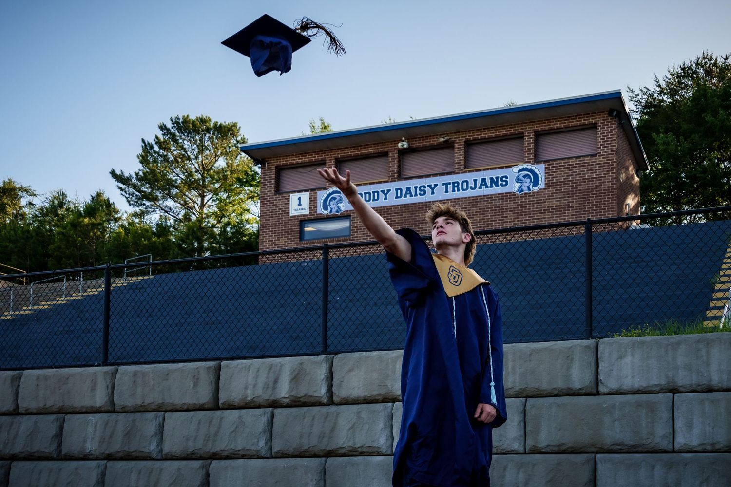 Graduate in blue cap and gown poses against stone wall during outdoor graduation photo session.