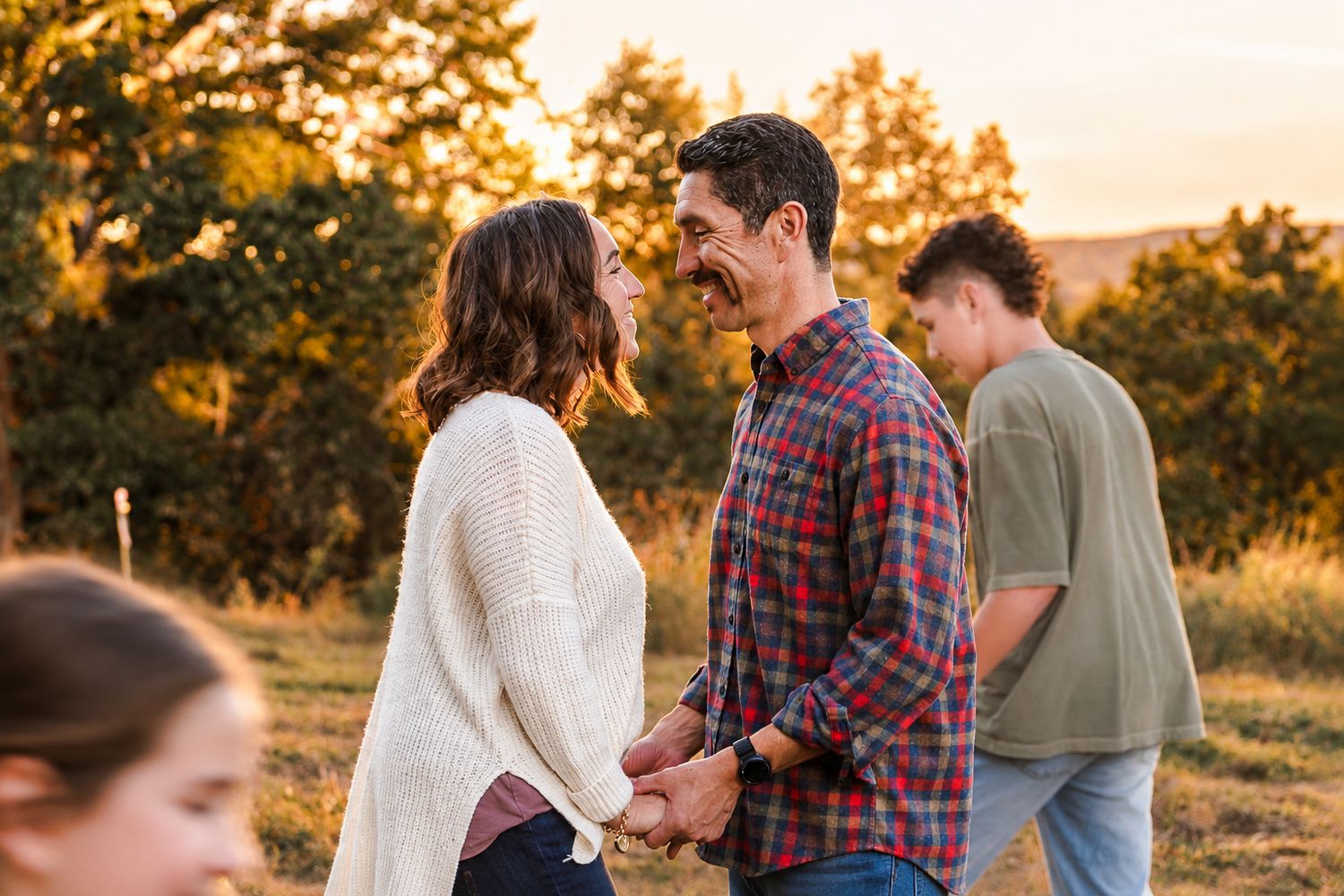A smiling couple holds hands and gazes at each other during golden hour in an outdoor field while a teen walks nearby and a child looks on.