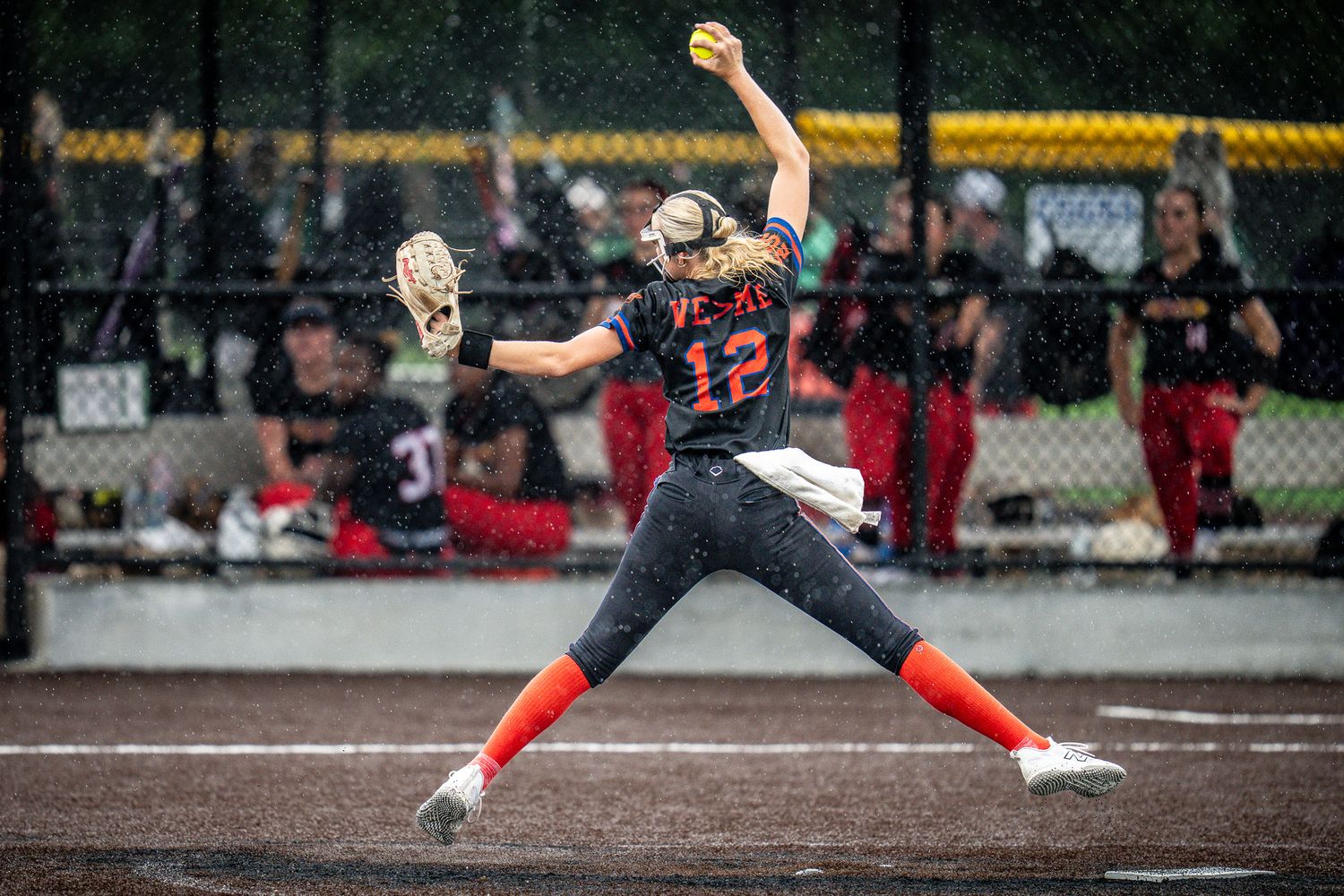 Softball pitcher #12 in orange uniform delivers a pitch in the rain during an intense game.