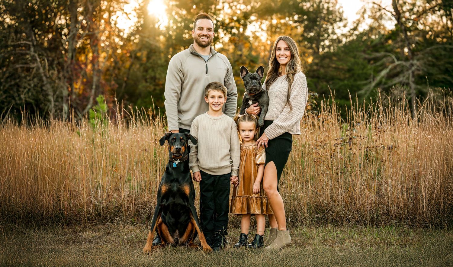 A family stands together in a golden field during sunset while their black dog sits beside them.