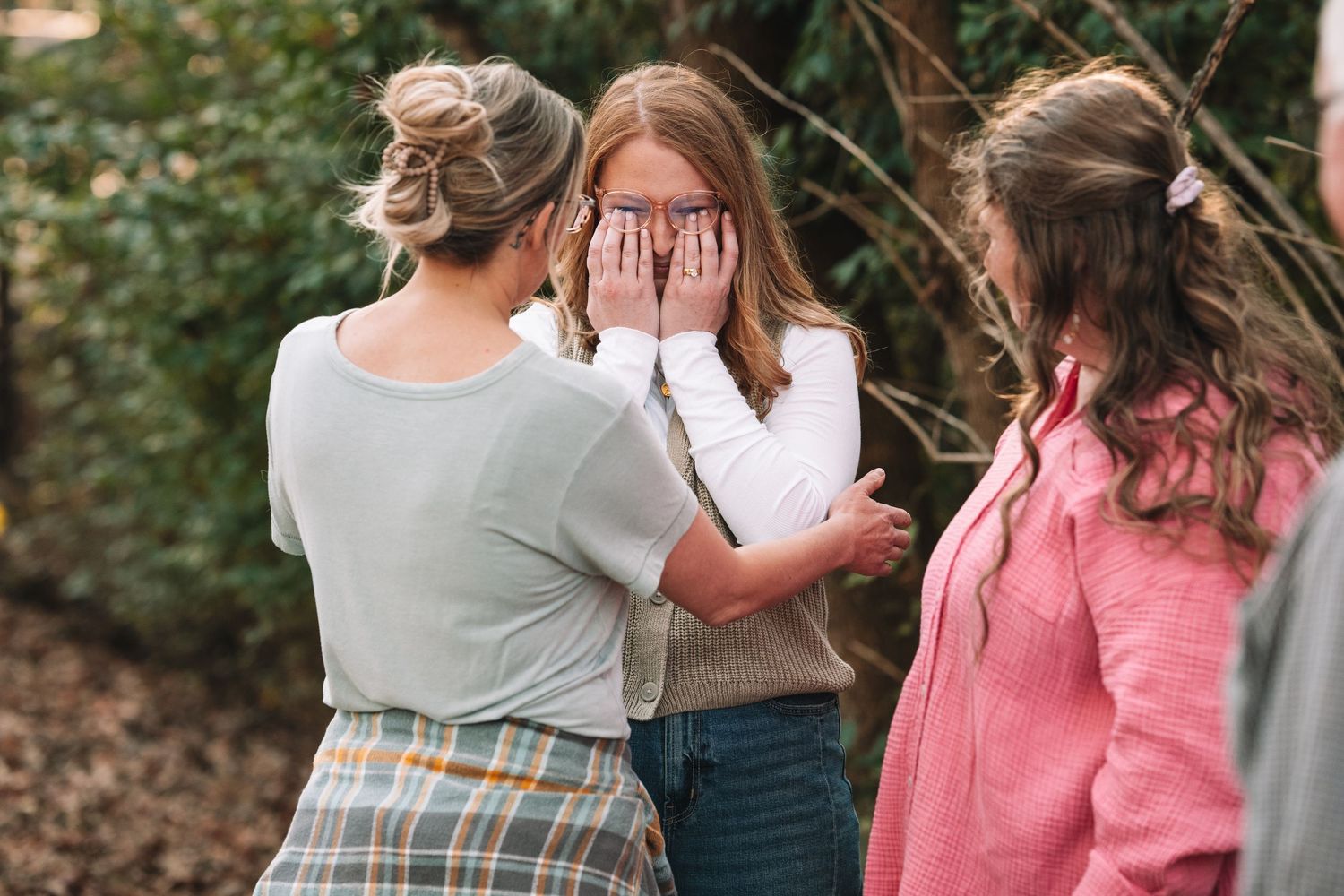 Family members embrace and laugh together while wearing coordinated casual outfits in an outdoor wooded setting.