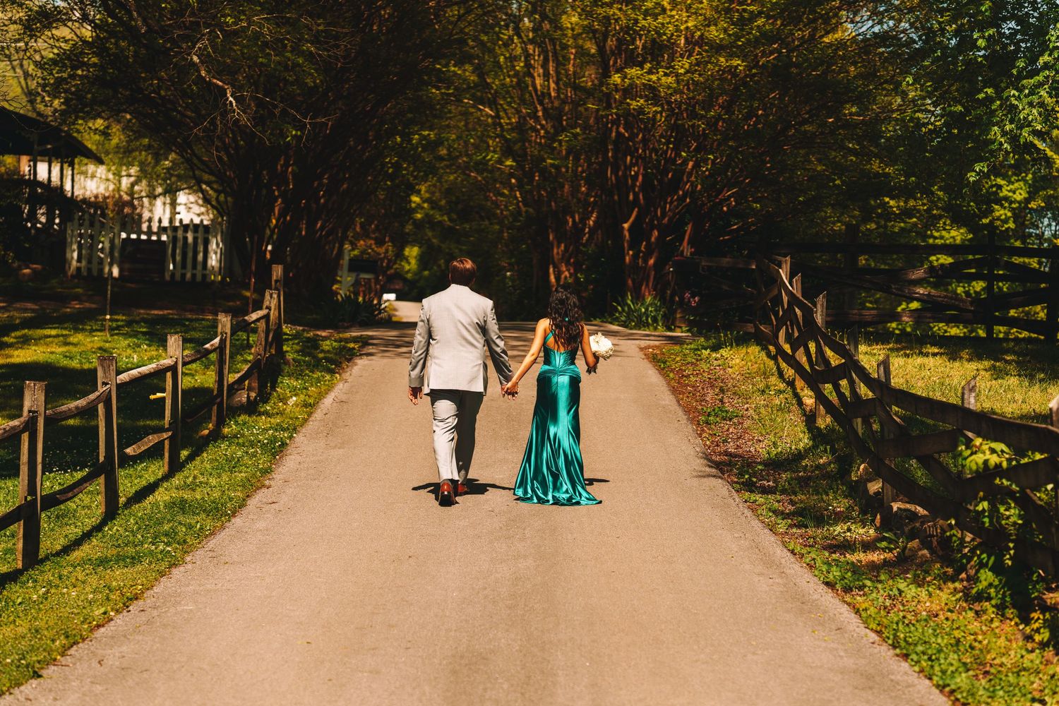 Couple in formal attire holding hands while walking down a tree-lined path flanked by wooden fences.