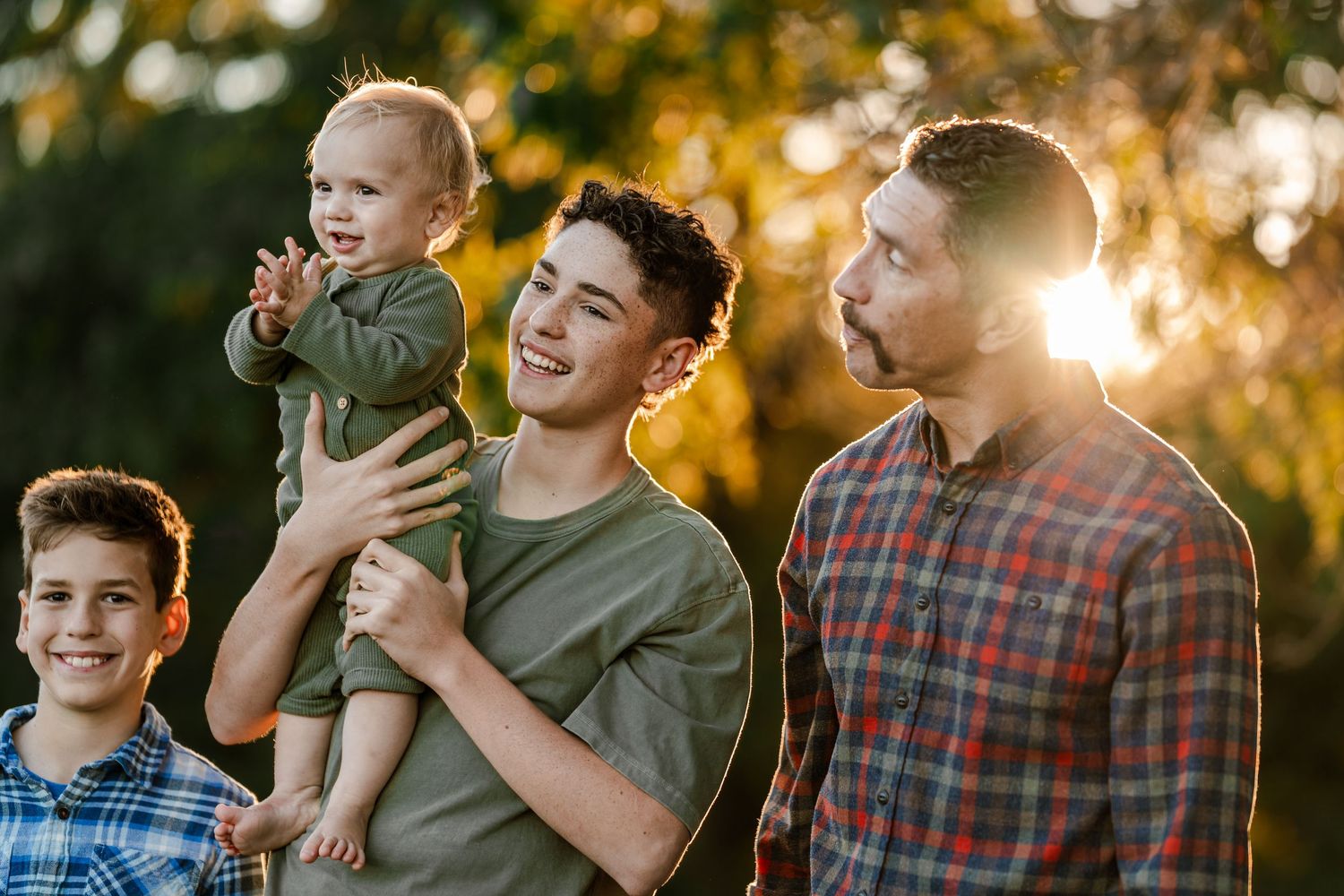 Smiling teenage boy holds a laughing baby beside a bearded man and a young boy outdoors in warm golden sunset light.