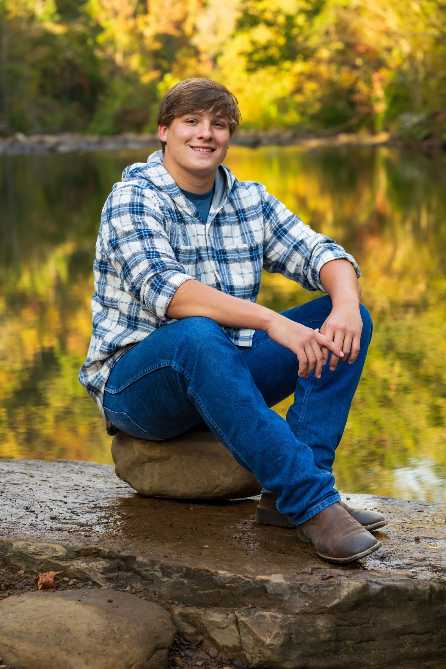 Person in plaid shirt and jeans sitting on rocks by a lake with autumn foliage reflections.