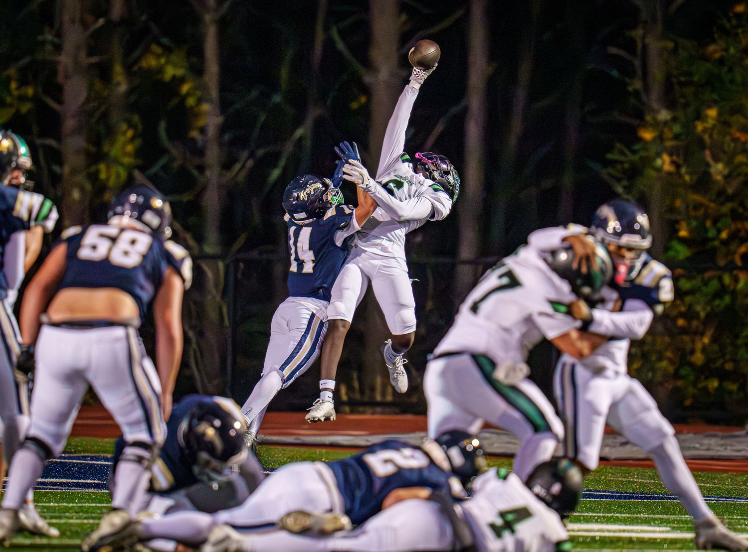Football player leaps to catch pass during night game with defenders and teammates on field.