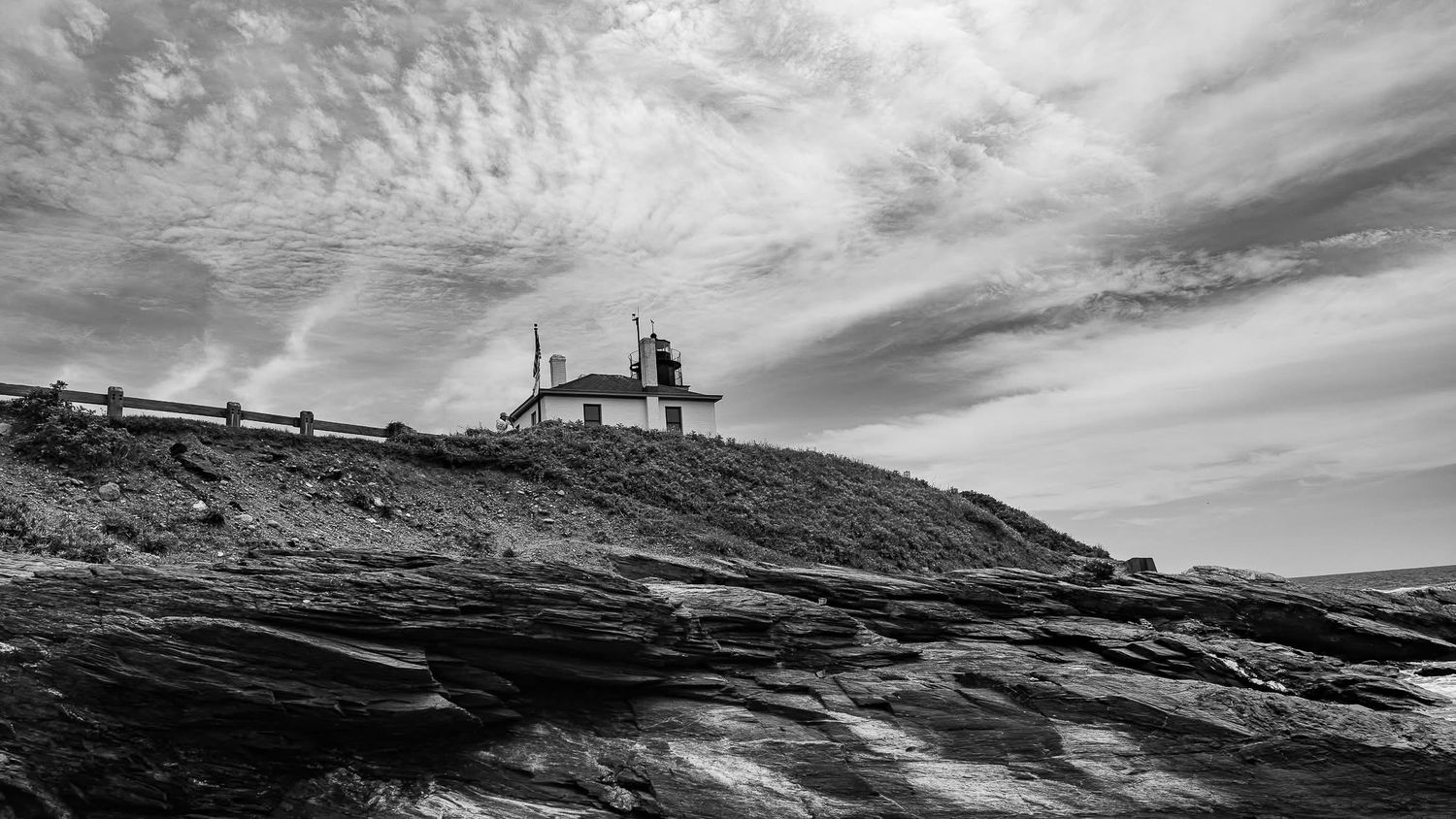 Black and white photo of a lighthouse perched on rocky coastal cliffs under a dramatic cloudy sky.