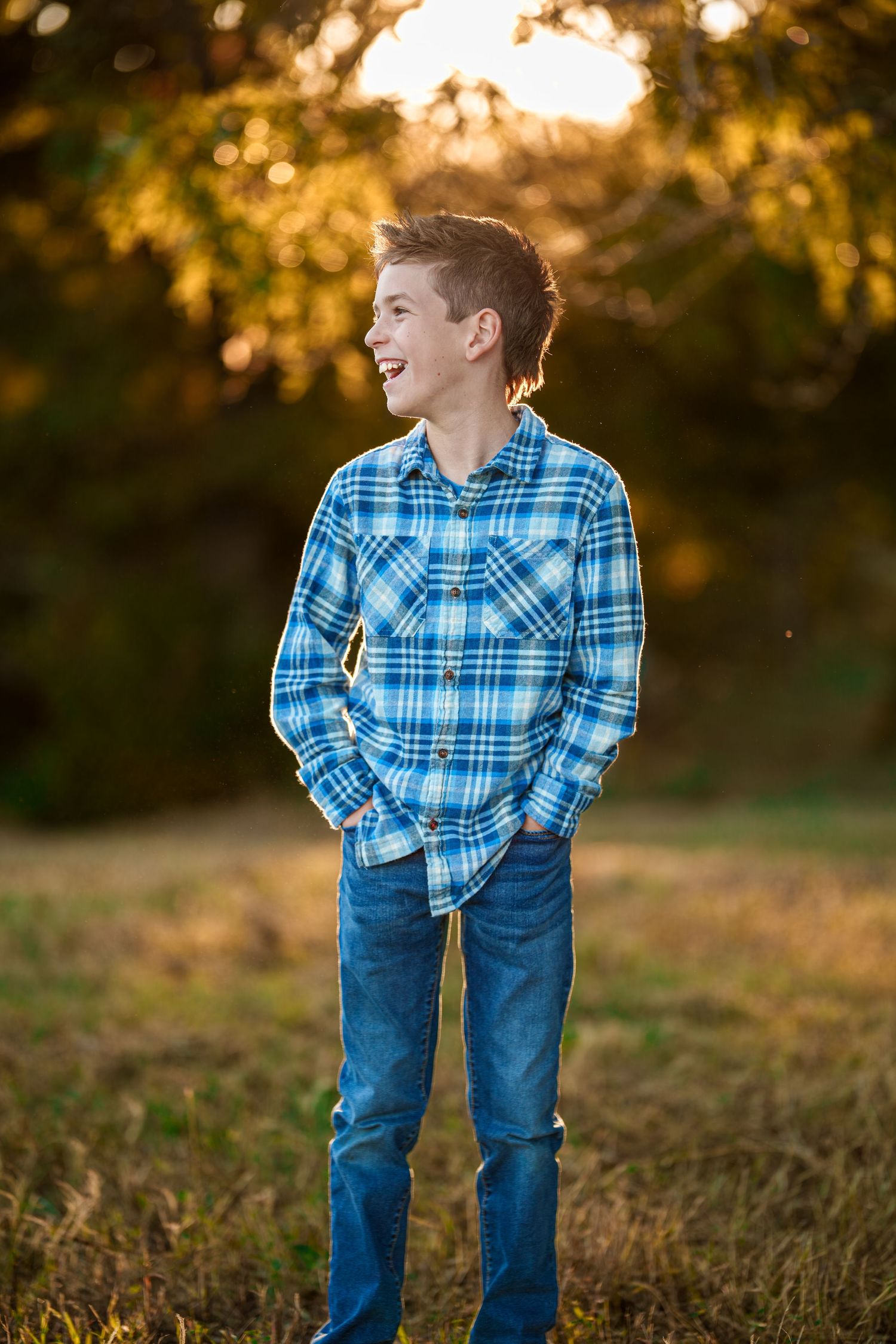Smiling boy in a blue plaid flannel shirt and jeans stands with hands in pockets in a grassy field at golden hour sunset.