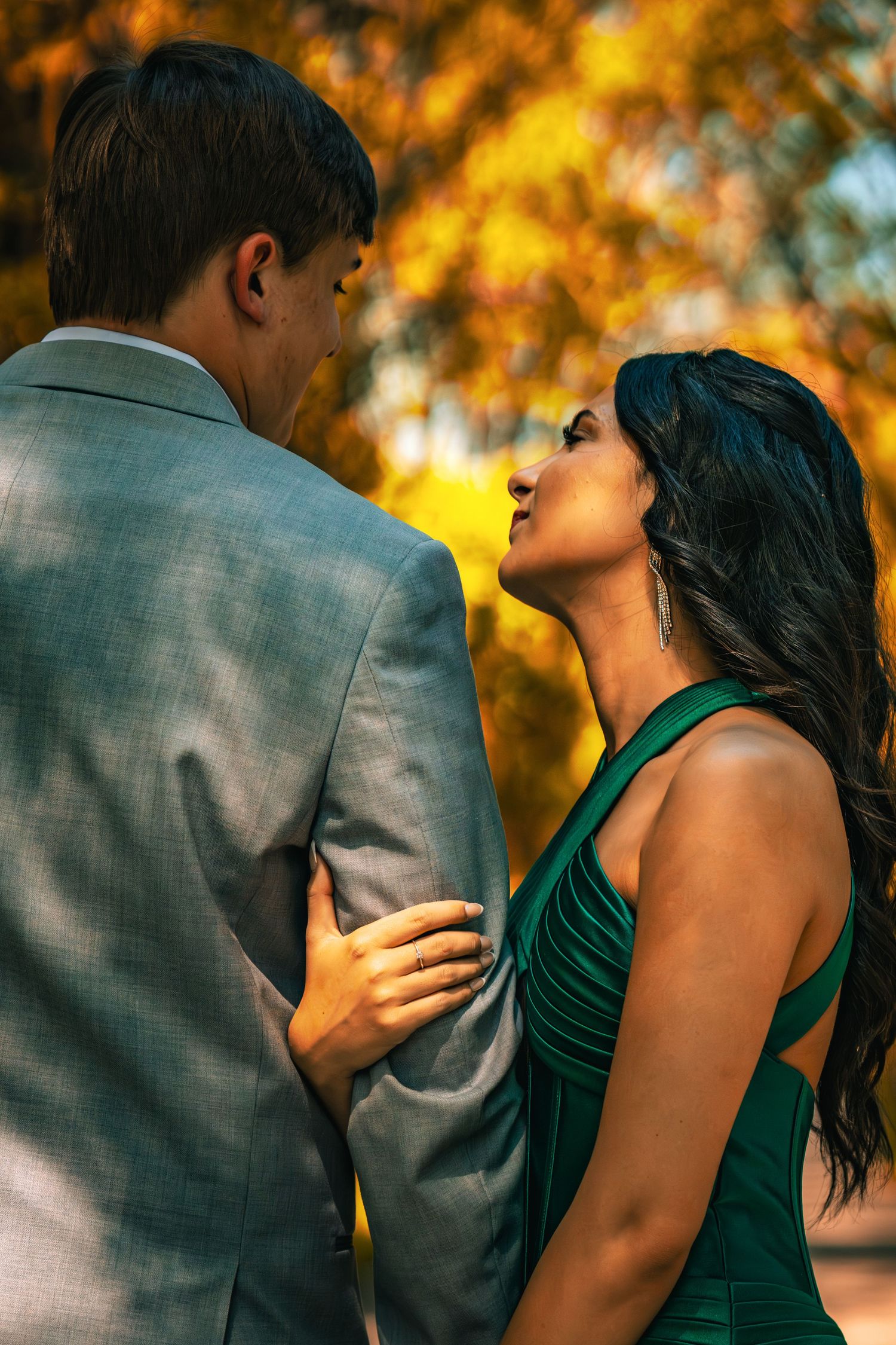 Couple in romantic moment outdoors, man in gray suit, woman in green dress, autumn foliage background.