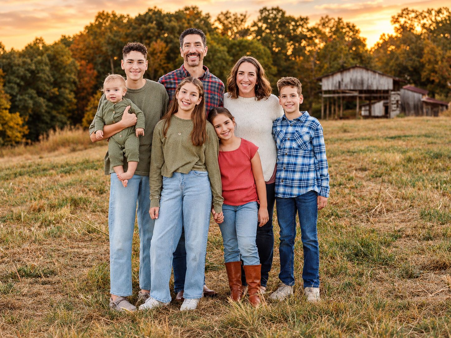 Smiling family of seven posing together in a grassy field at sunset, with autumn trees and a rustic barn in the background.