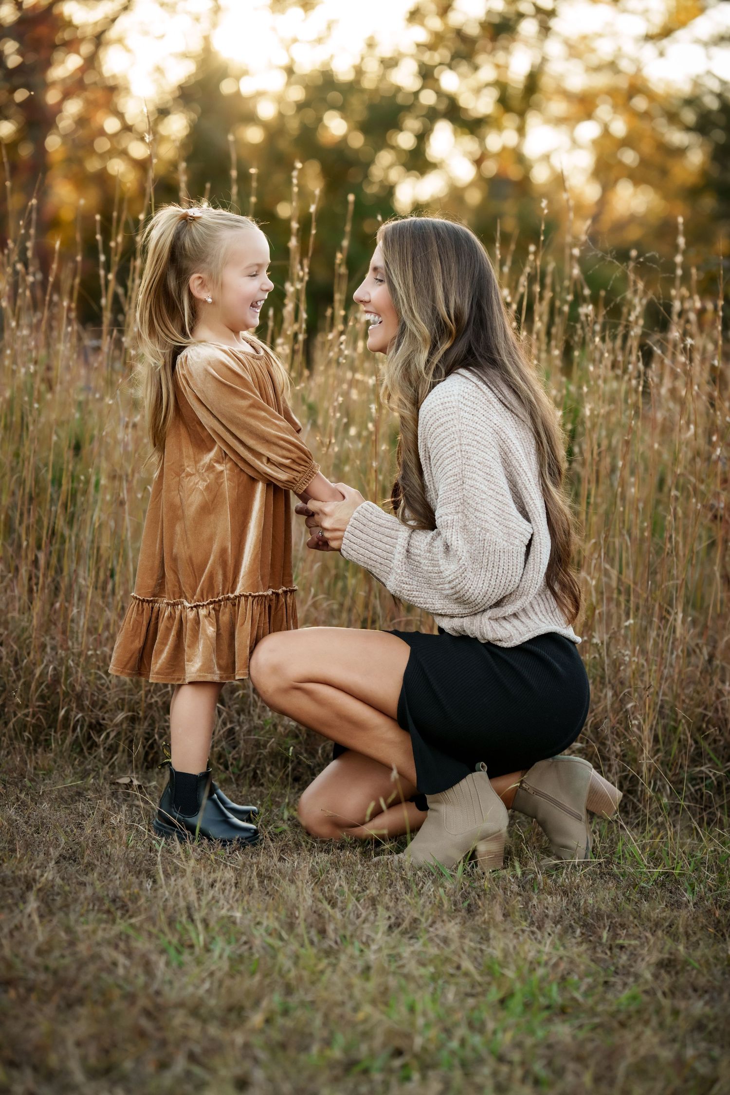 Two people sit together in a grassy field at sunset wearing cozy fall outfits and sharing a warm moment.