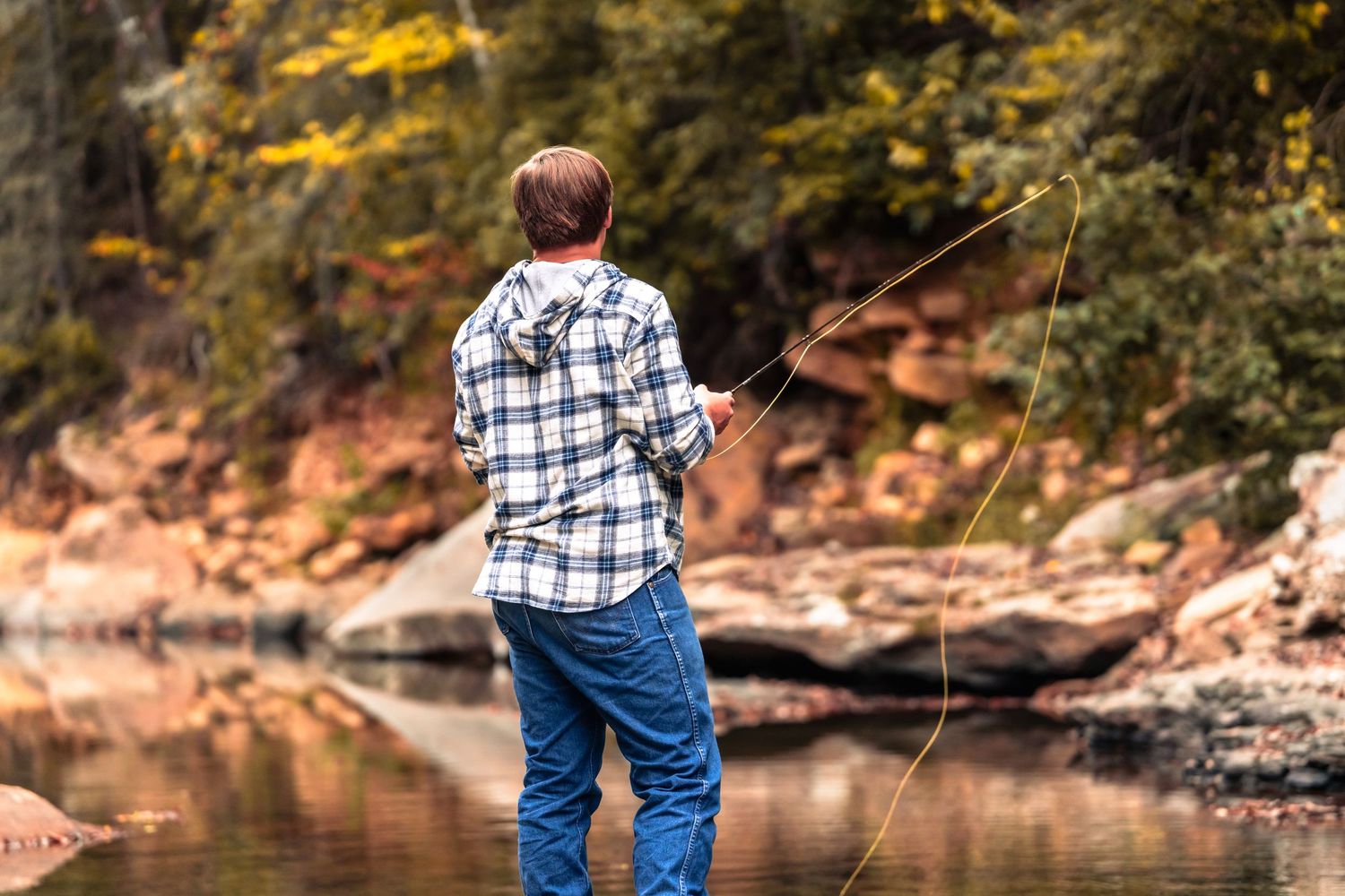 Person in plaid shirt fishing at scenic autumn riverbank.