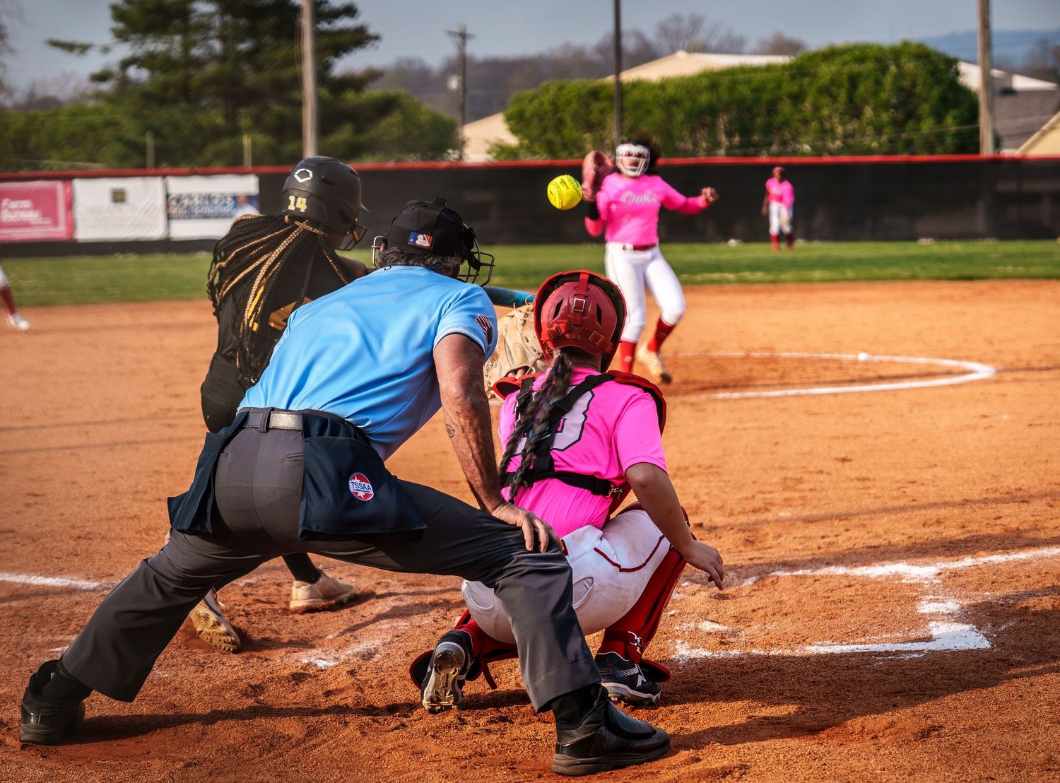 Softball game action shot with catcher in pink gear, umpire, and pitcher throwing ball on dirt field.