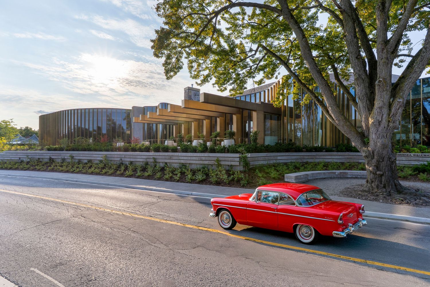 Classic red vintage car parked along a tree-lined street near modern buildings on a sunny day.