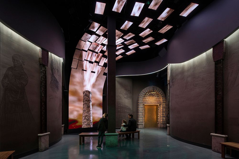 Immersive museum exhibition hall with dramatic lighting, large video displays, and ancient architectural elements on dark walls.