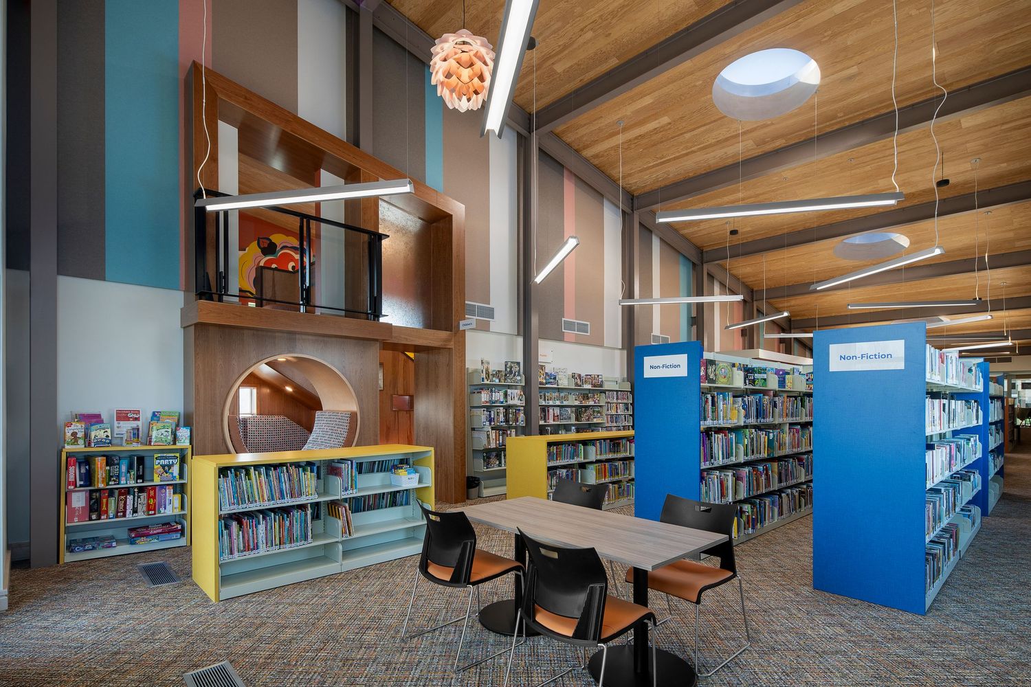 Modern library interior with colorful bookshelves, reading tables, wooden ceiling, and a children's section with bright displays.