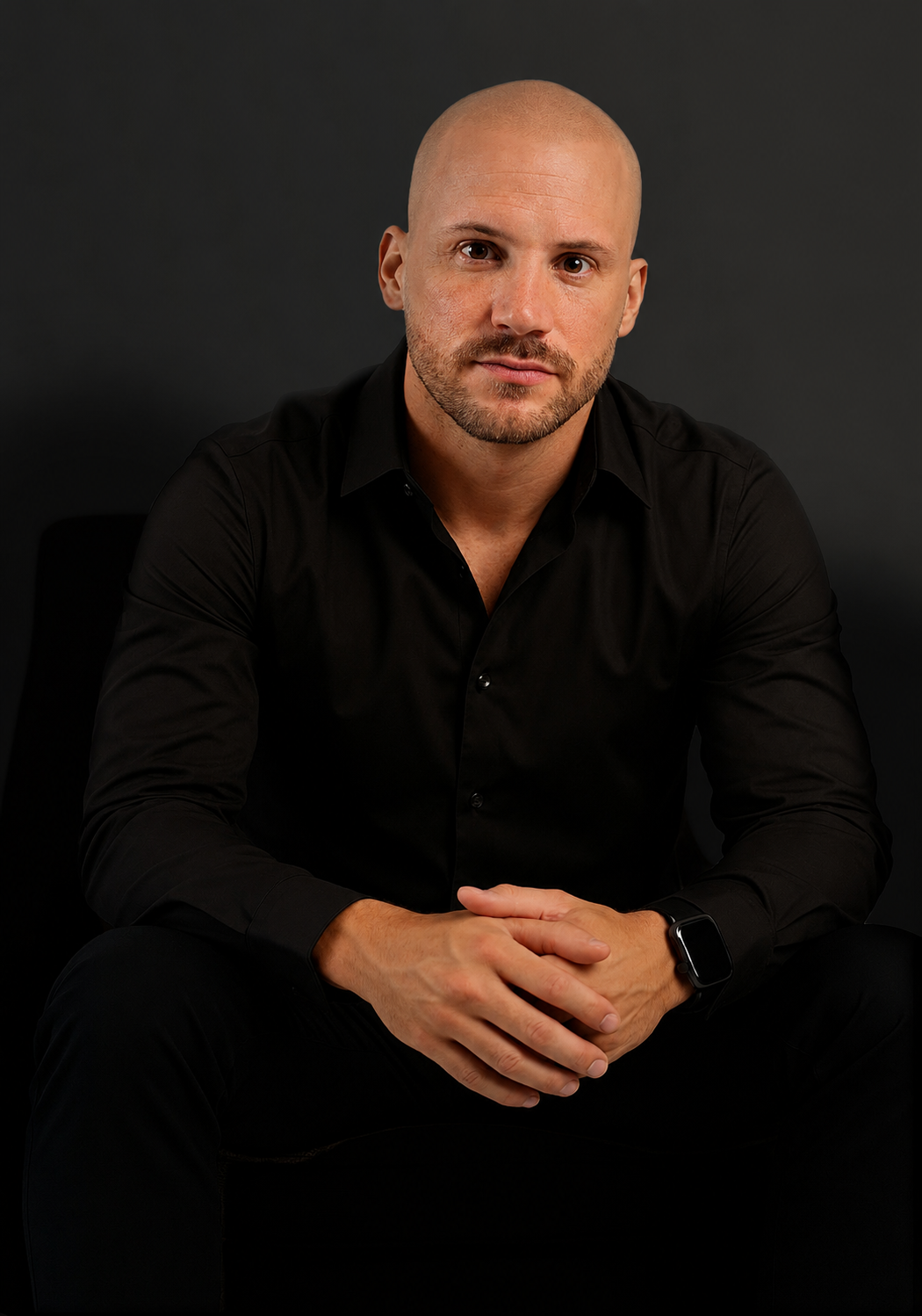 Bald man in black shirt sitting with hands clasped, posing confidently against dark background.