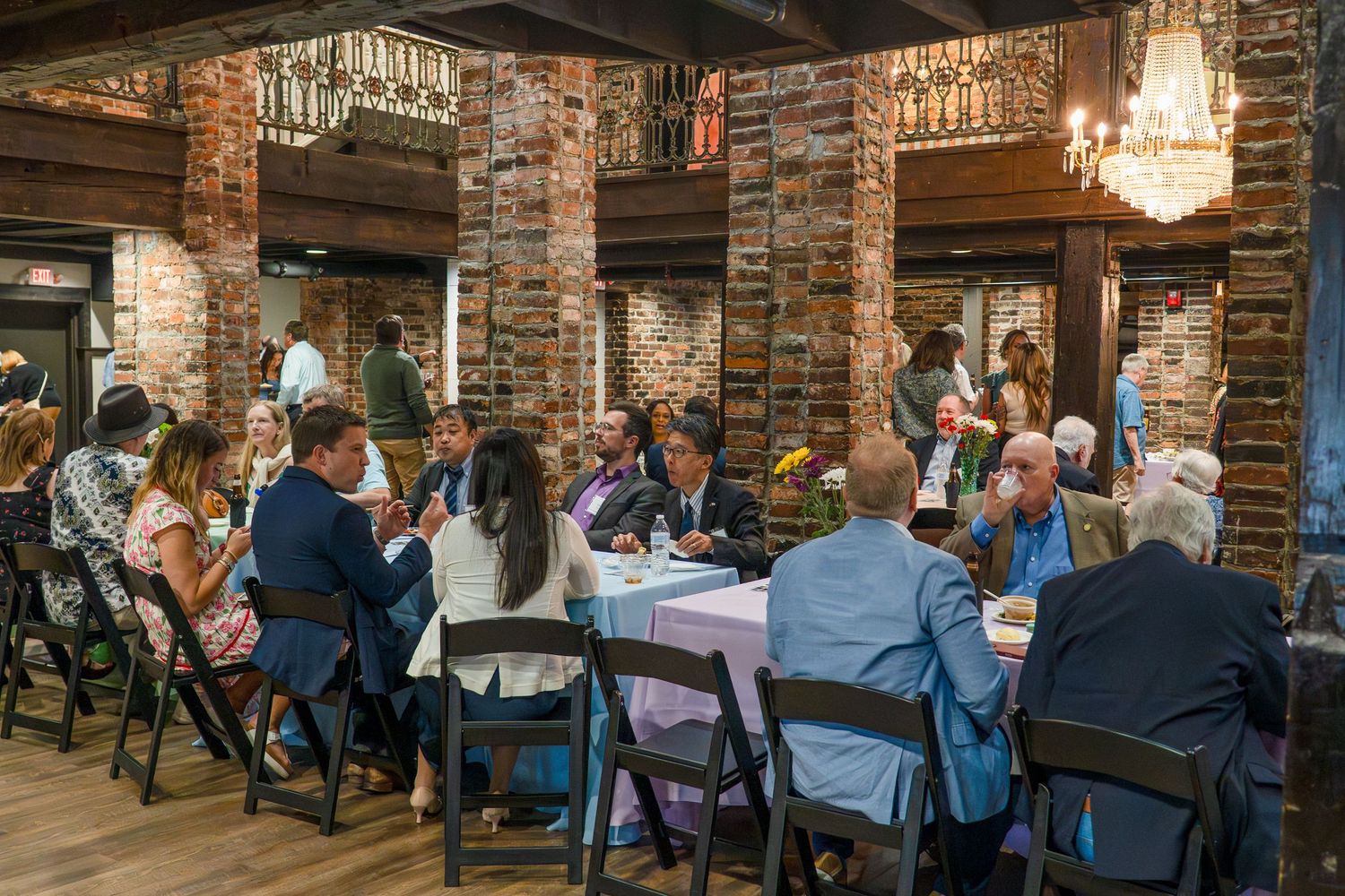 Elegant chandelier lighting illuminates tables of guests in a historic brick dining space.