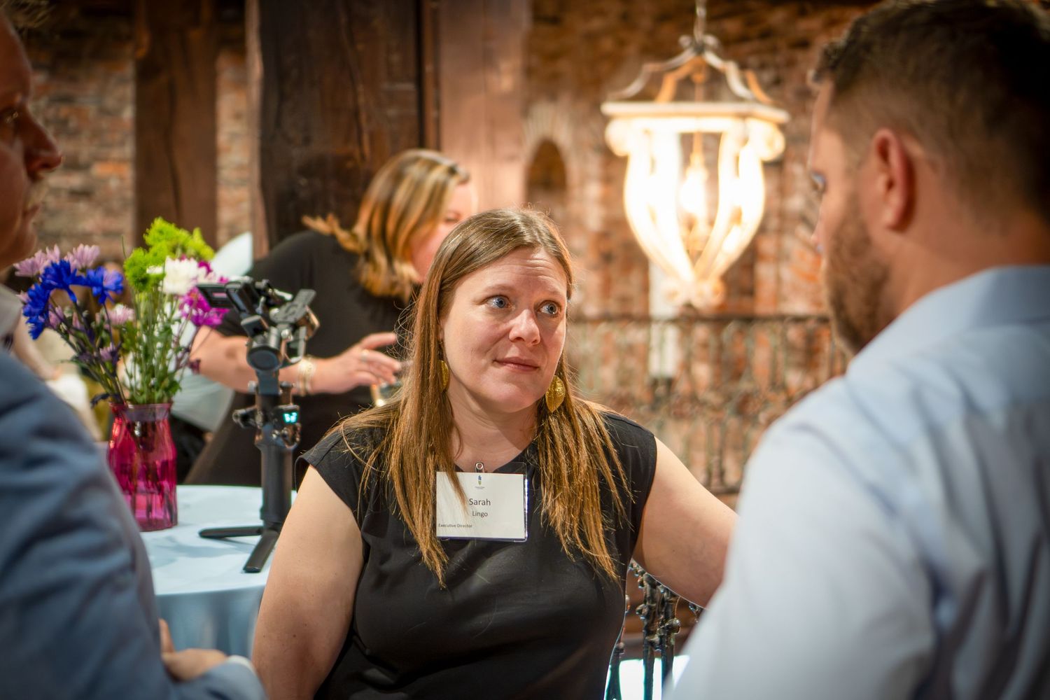 Guests engaged in conversation at an indoor event with brick walls and chandeliers.