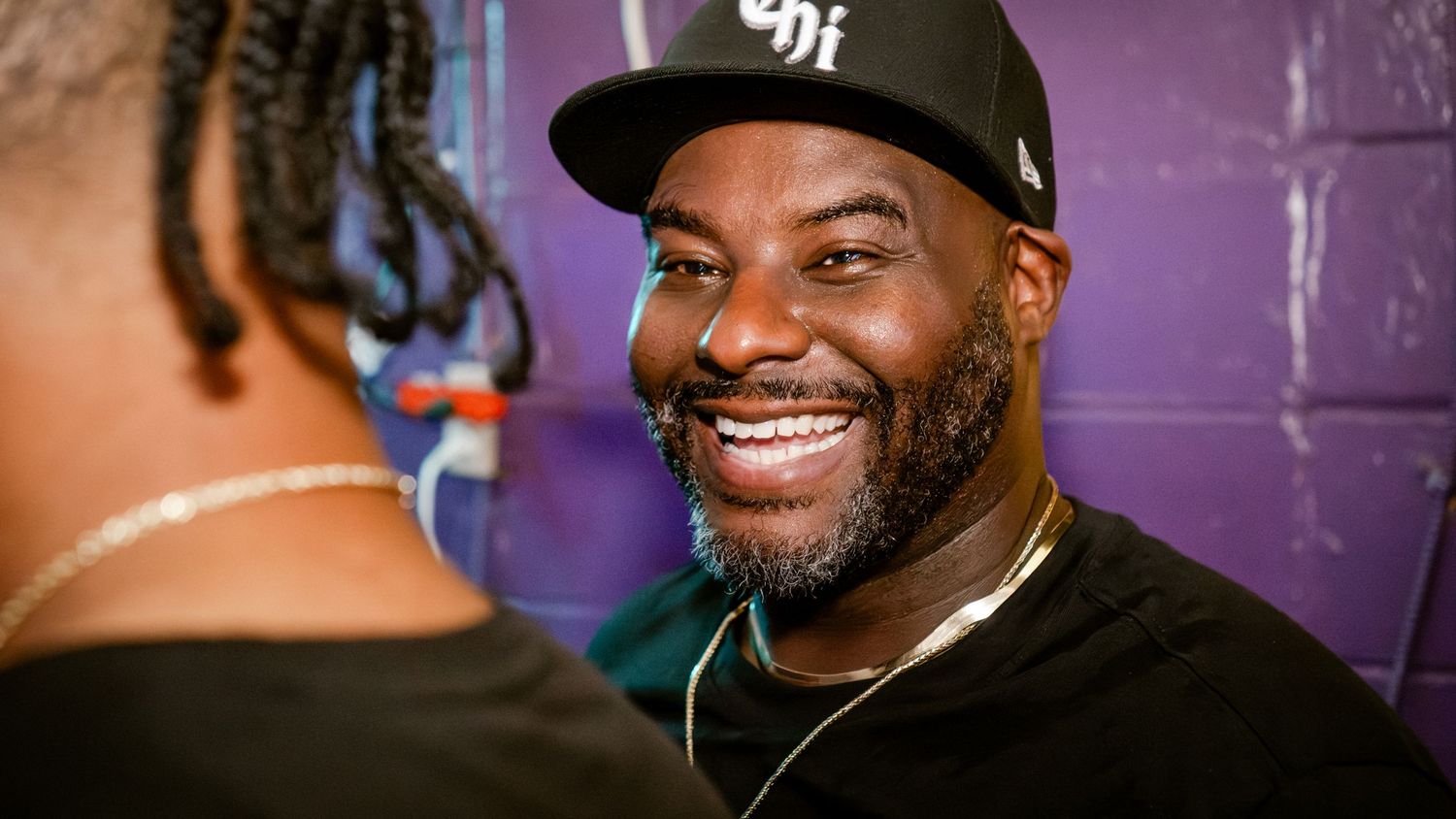 Man in baseball cap smiling against purple wall background.