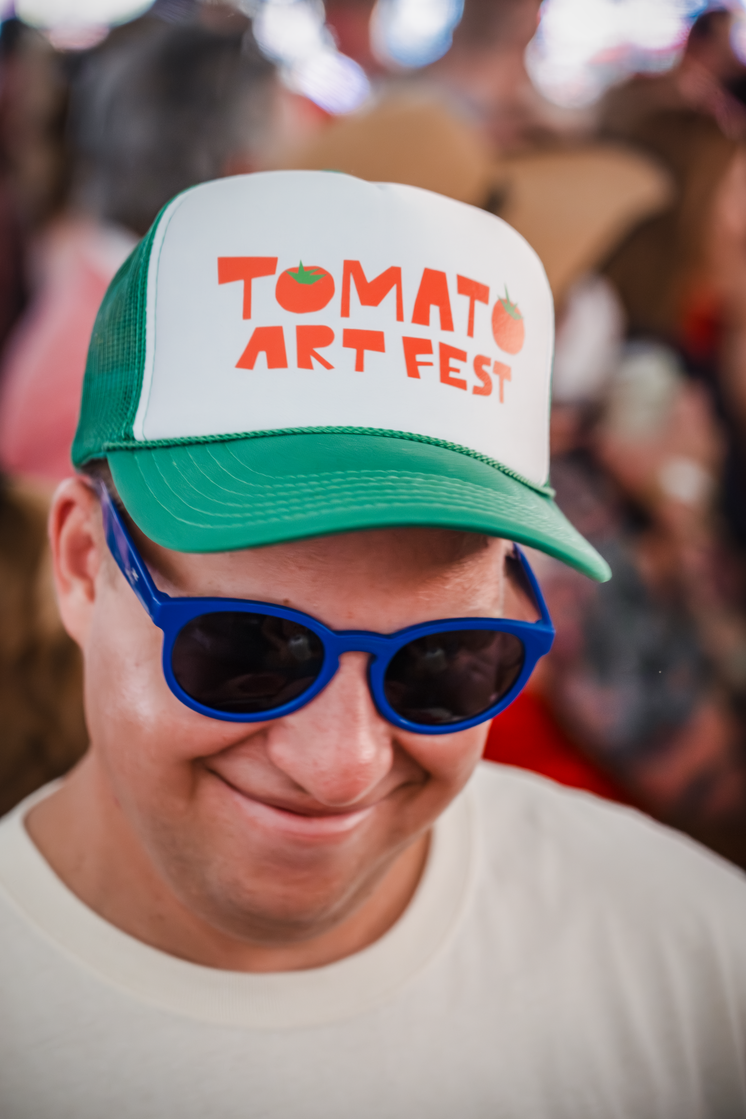 White baseball cap with green brim and blue sunglasses displaying Tomato Art Fest branding.