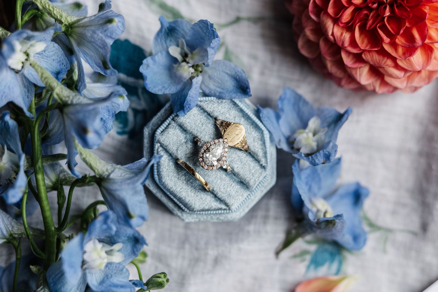 Gold engagement ring with diamond in a blue velvet ring box surrounded by blue delphinium flowers and a pink dahlia.