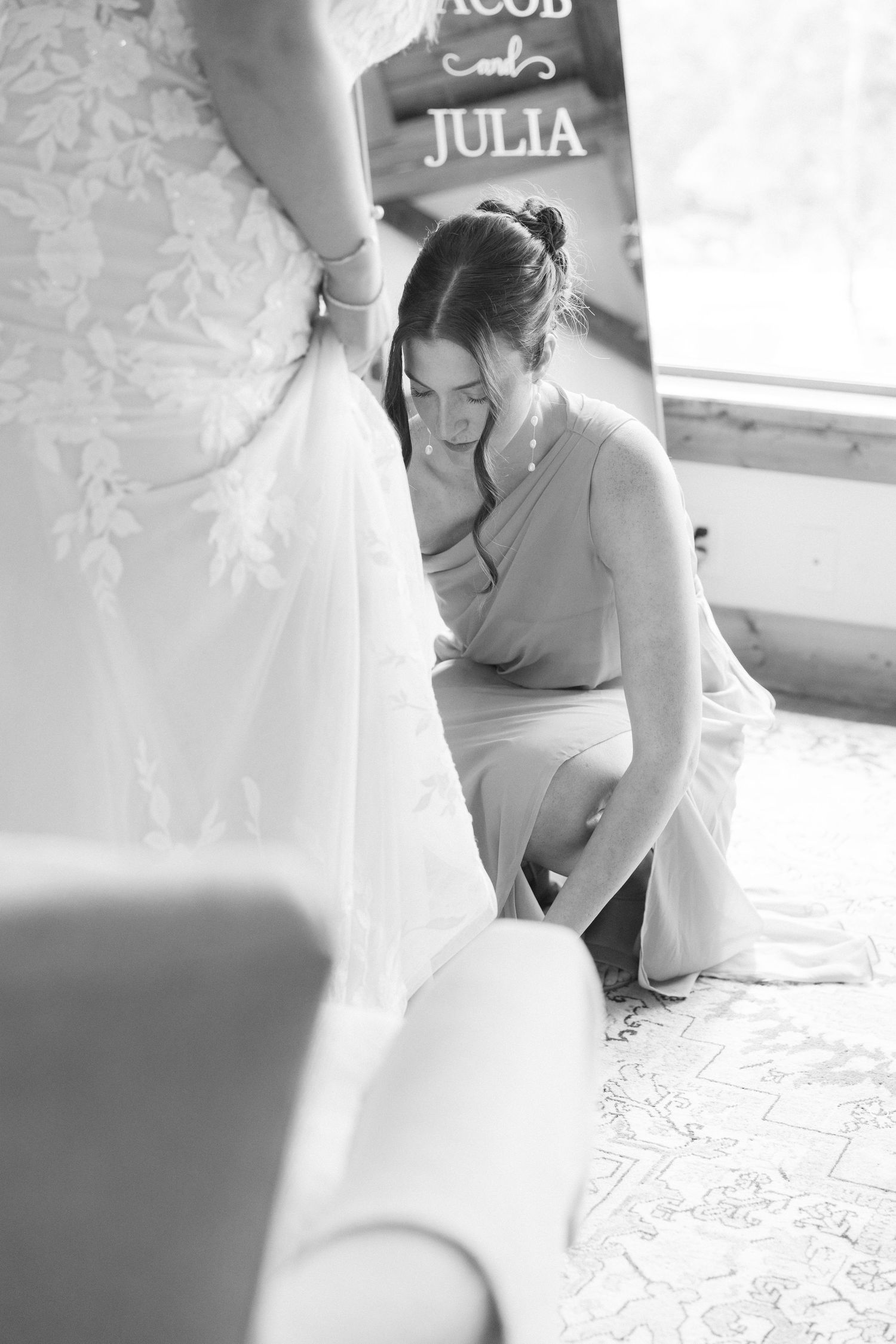 Bridesmaid kneeling to adjust brides wedding gown hem in black and white bridal preparation photo.